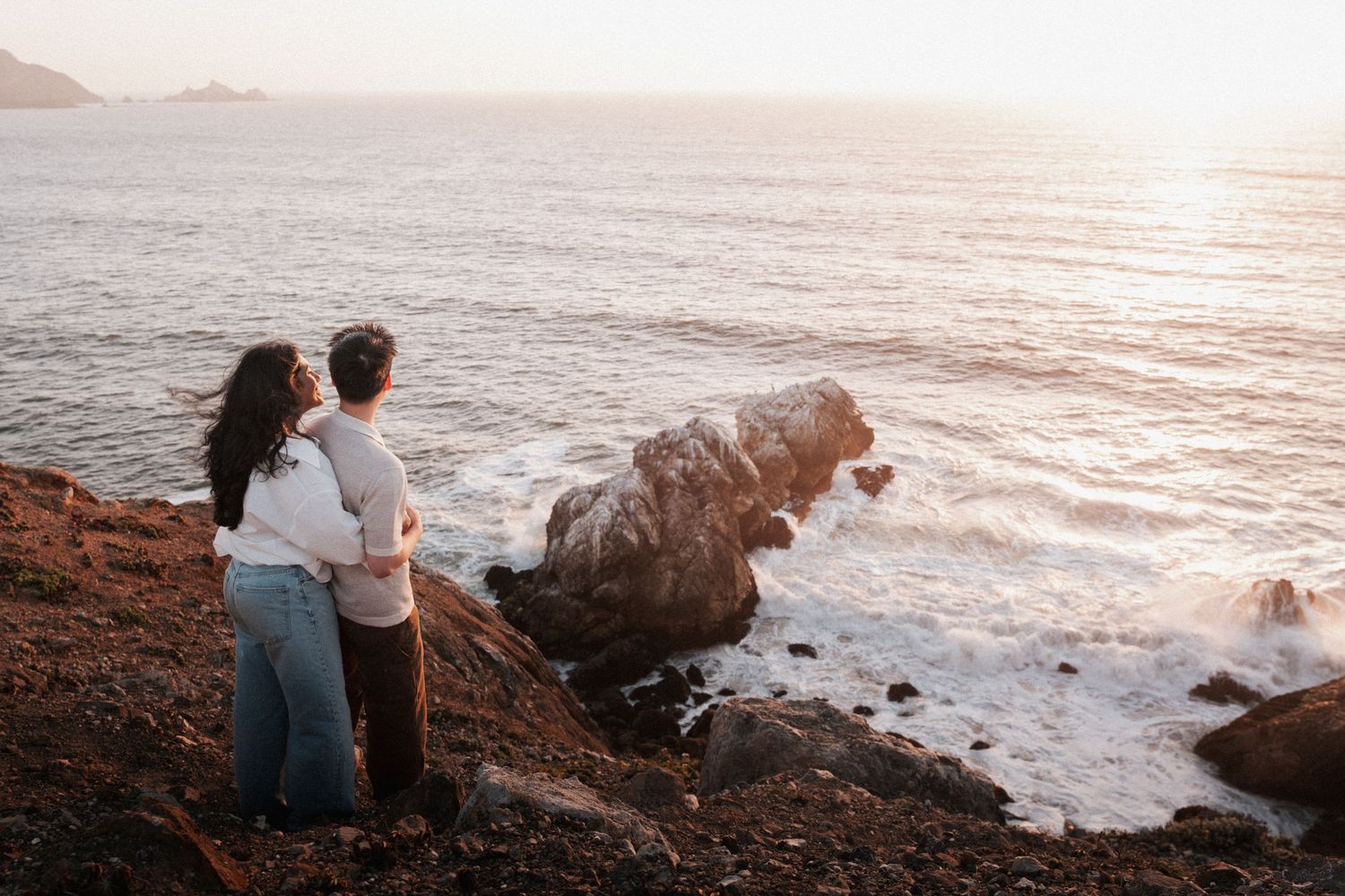 Couple embracing on rocky coastal cliffs watching a serene sunset over the ocean waves.