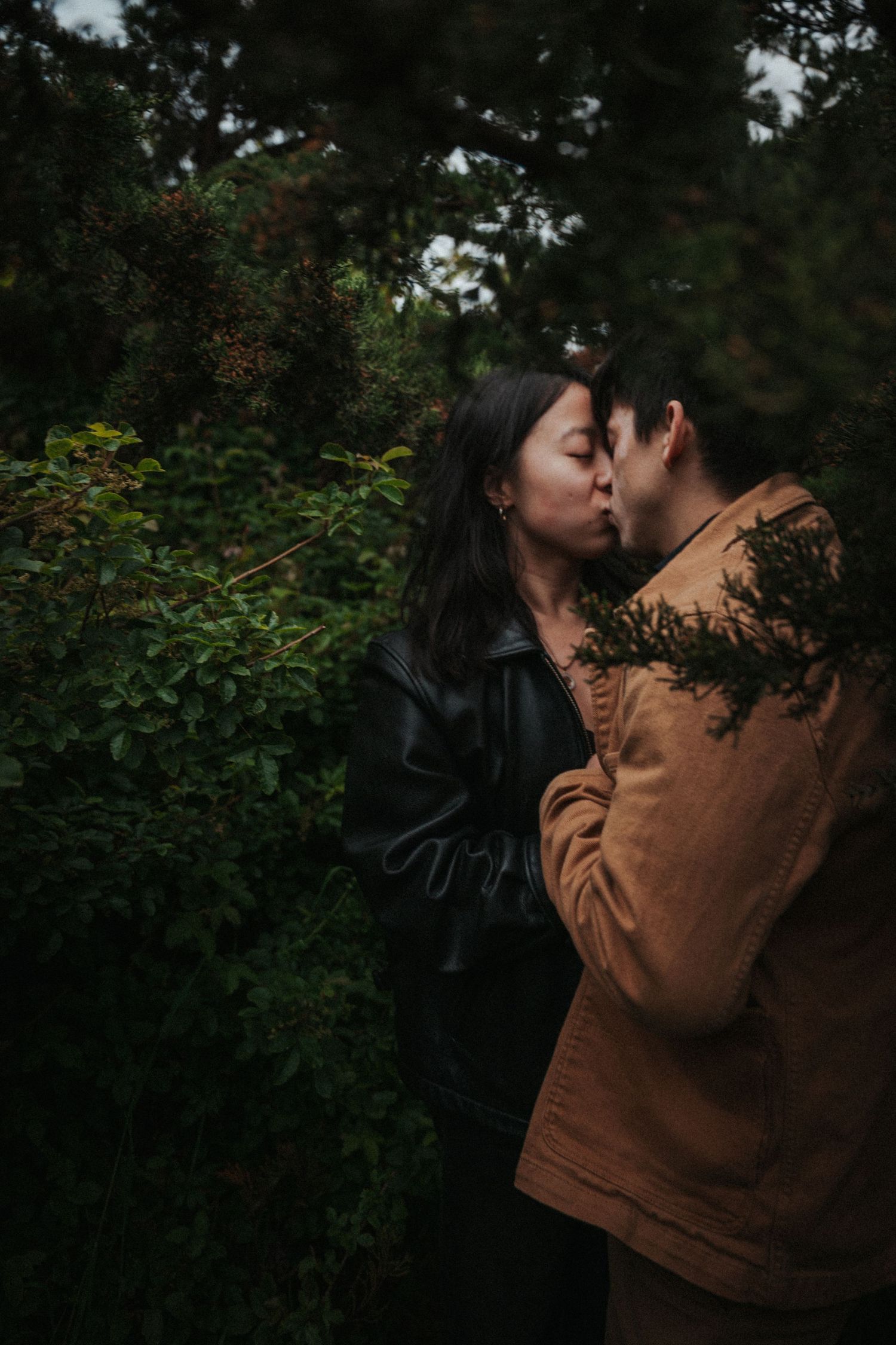 Couple kissing tenderly among lush green trees wearing leather and suede jackets.