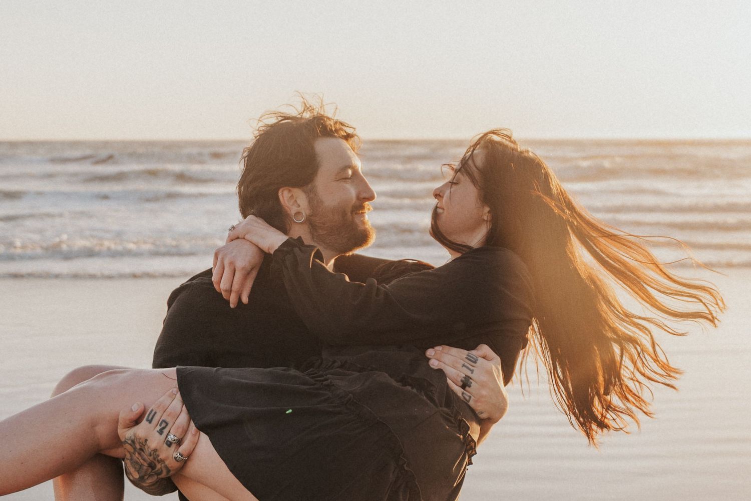 Romantic couple embracing on a beach at sunset, man holding woman in his arms near ocean waves.