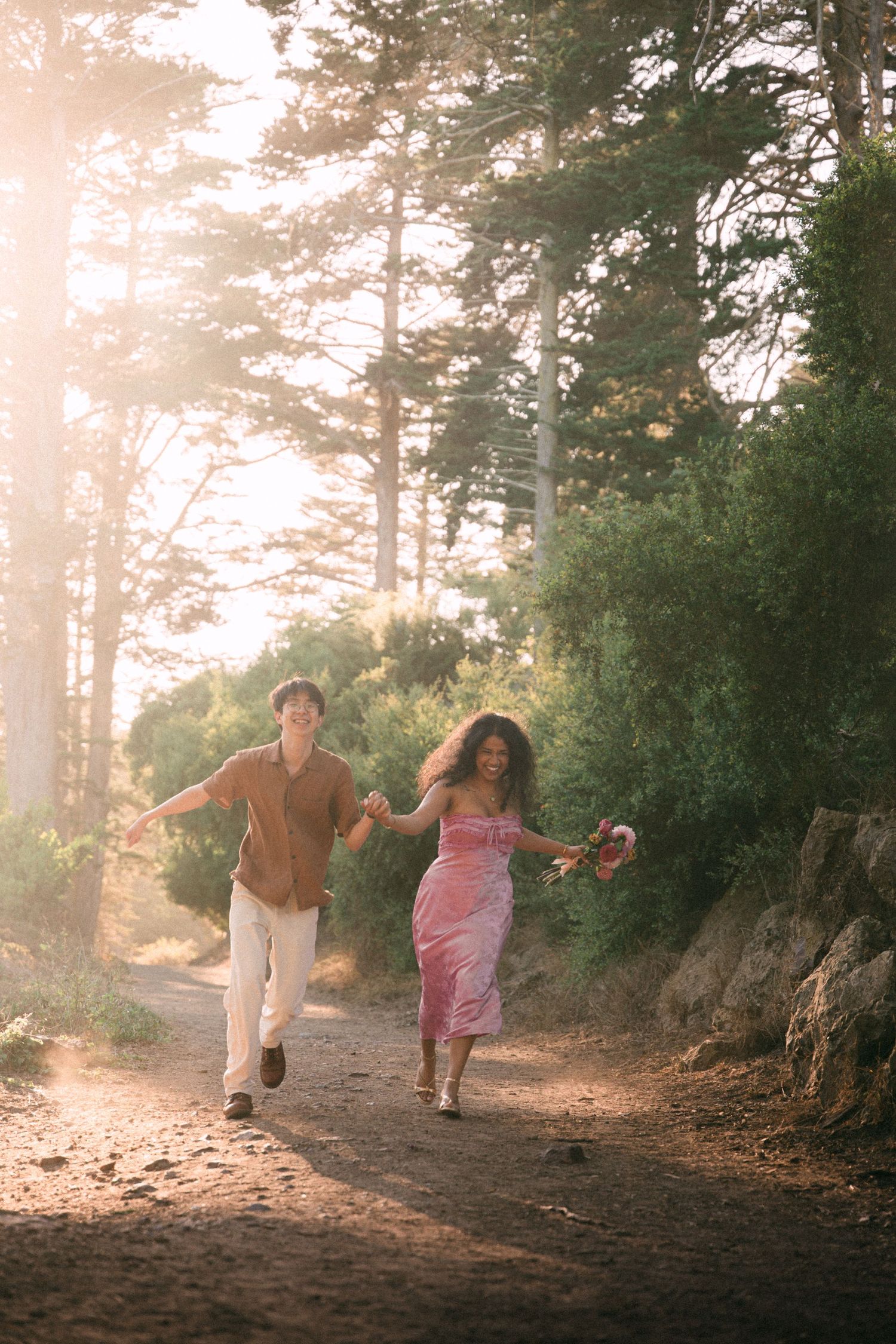 A joyful couple runs hand-in-hand along a sunlit forest path, woman in pink dress holding flowers.