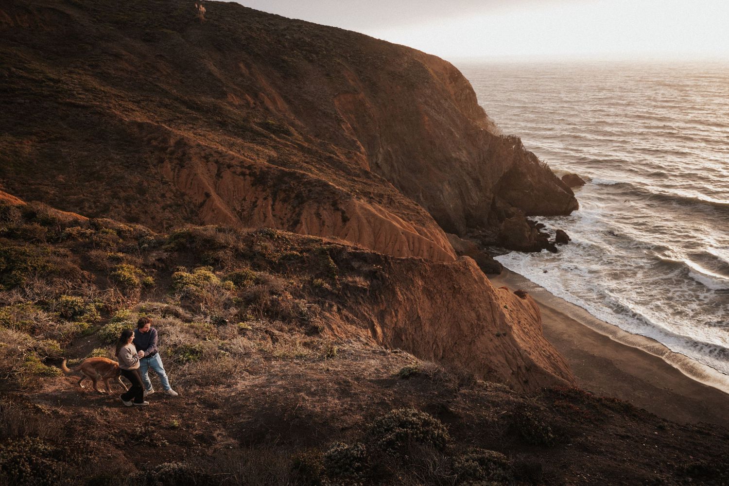 Couple standing on rugged coastal cliffs at golden hour with ocean waves below.