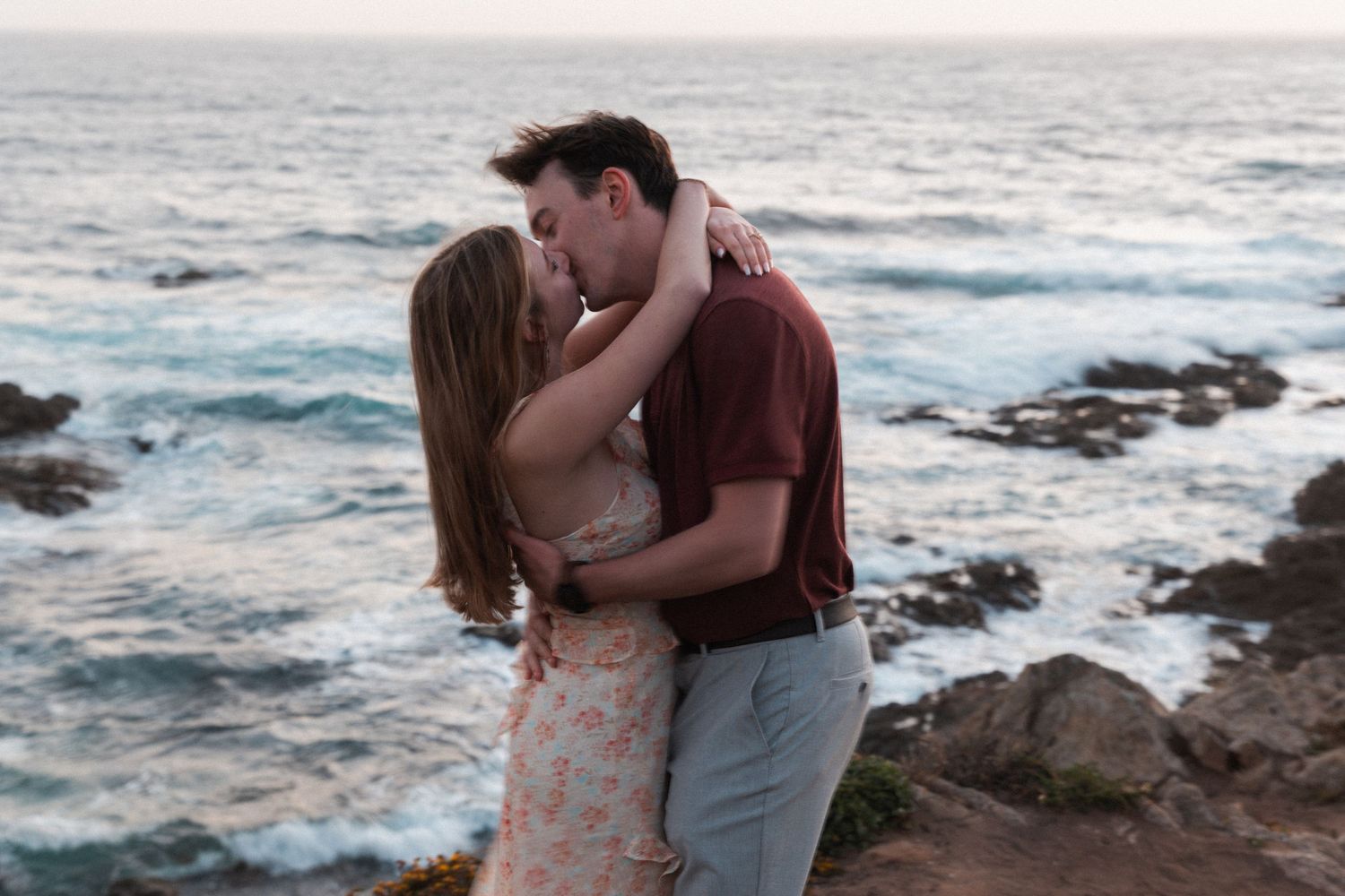 Couple sharing a romantic kiss on rocky coastal cliffs with ocean waves in the background at sunset.