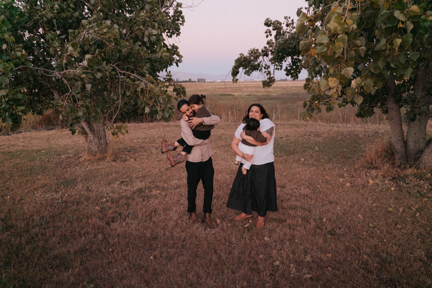 Two couples embrace outdoors among trees in a golden field at dusk, one pair hugging and another holding a baby.