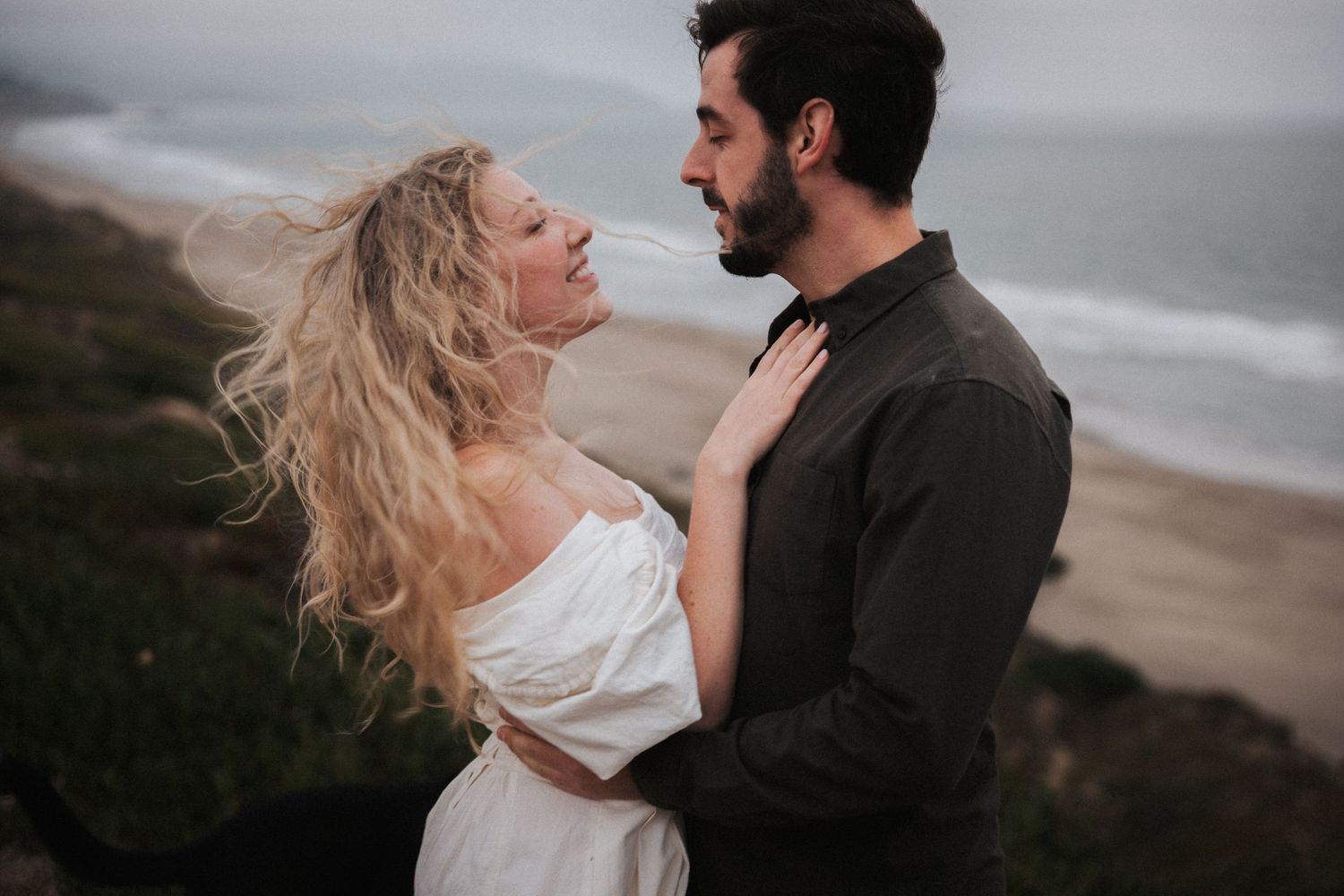 Couple embracing on a coastal cliff, woman in white off-shoulder dress with windswept blonde hair, man in dark shirt.
