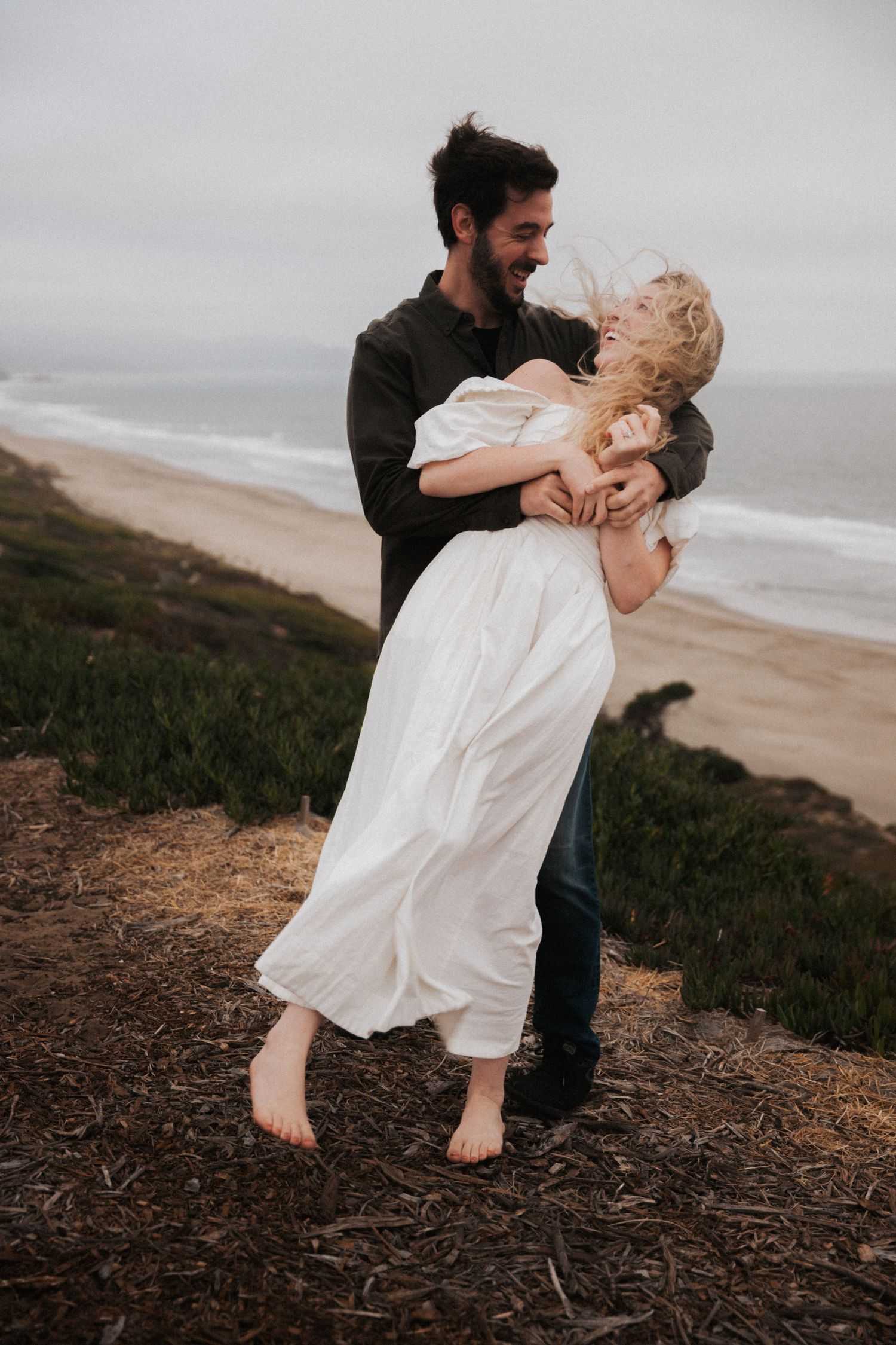 A laughing couple embraces on a coastal bluff, the man lifting the woman in a white dress.