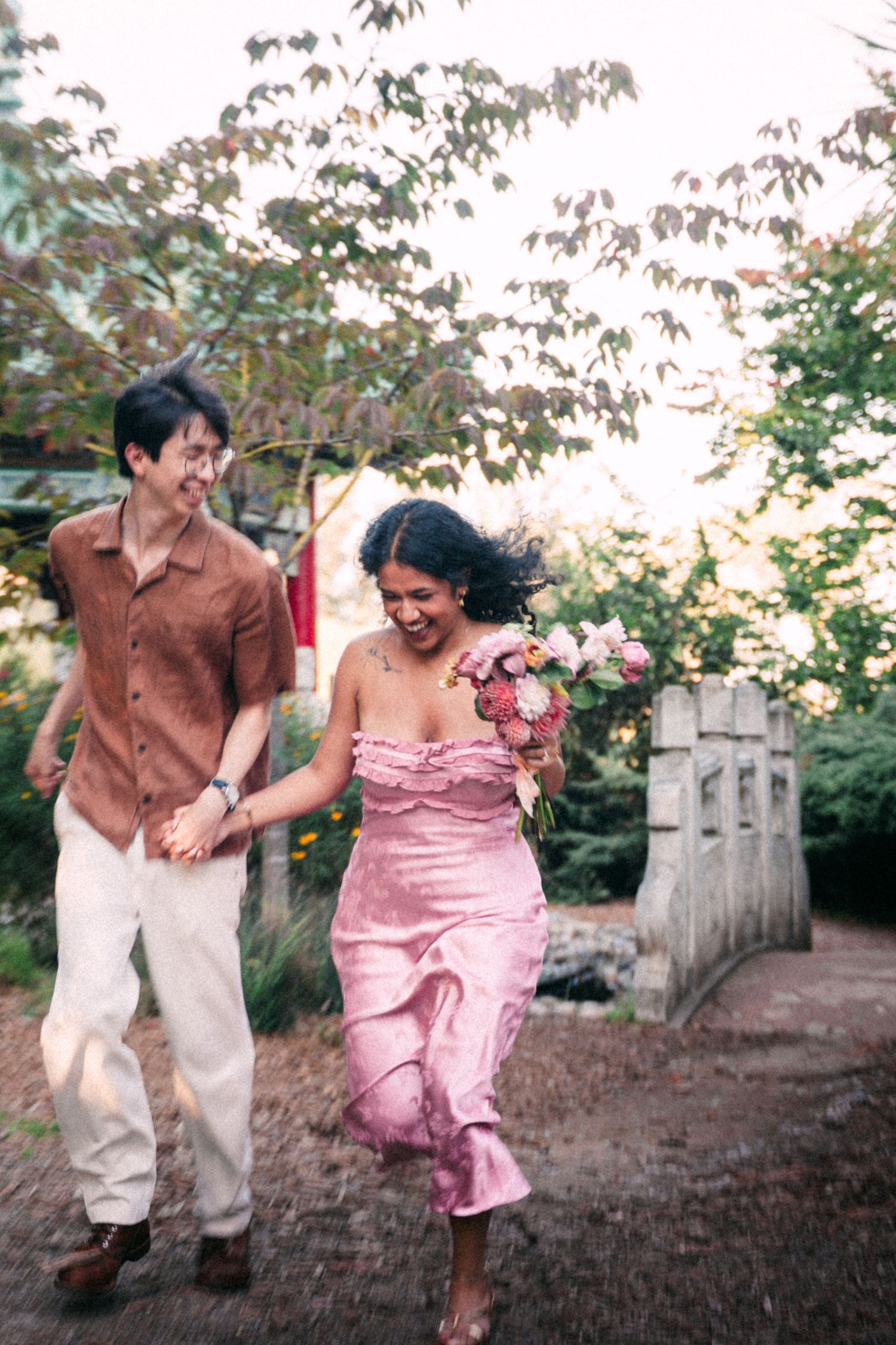 A joyful couple runs hand-in-hand outdoors, woman in pink strapless dress holding bouquet, man in brown shirt and white pants.