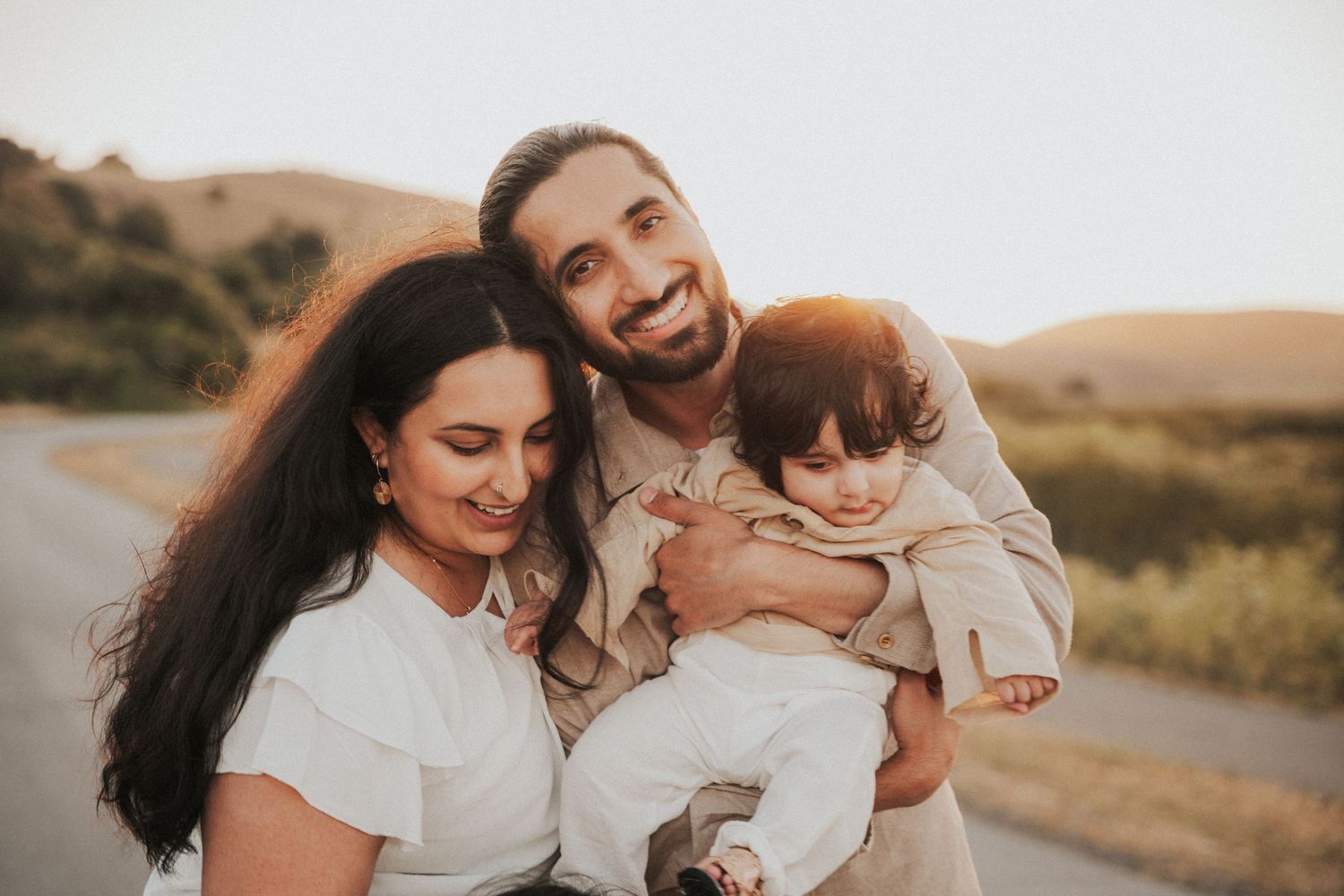 Happy young family with bearded father and dark-haired mother smiling while holding baby outdoors at sunset.
