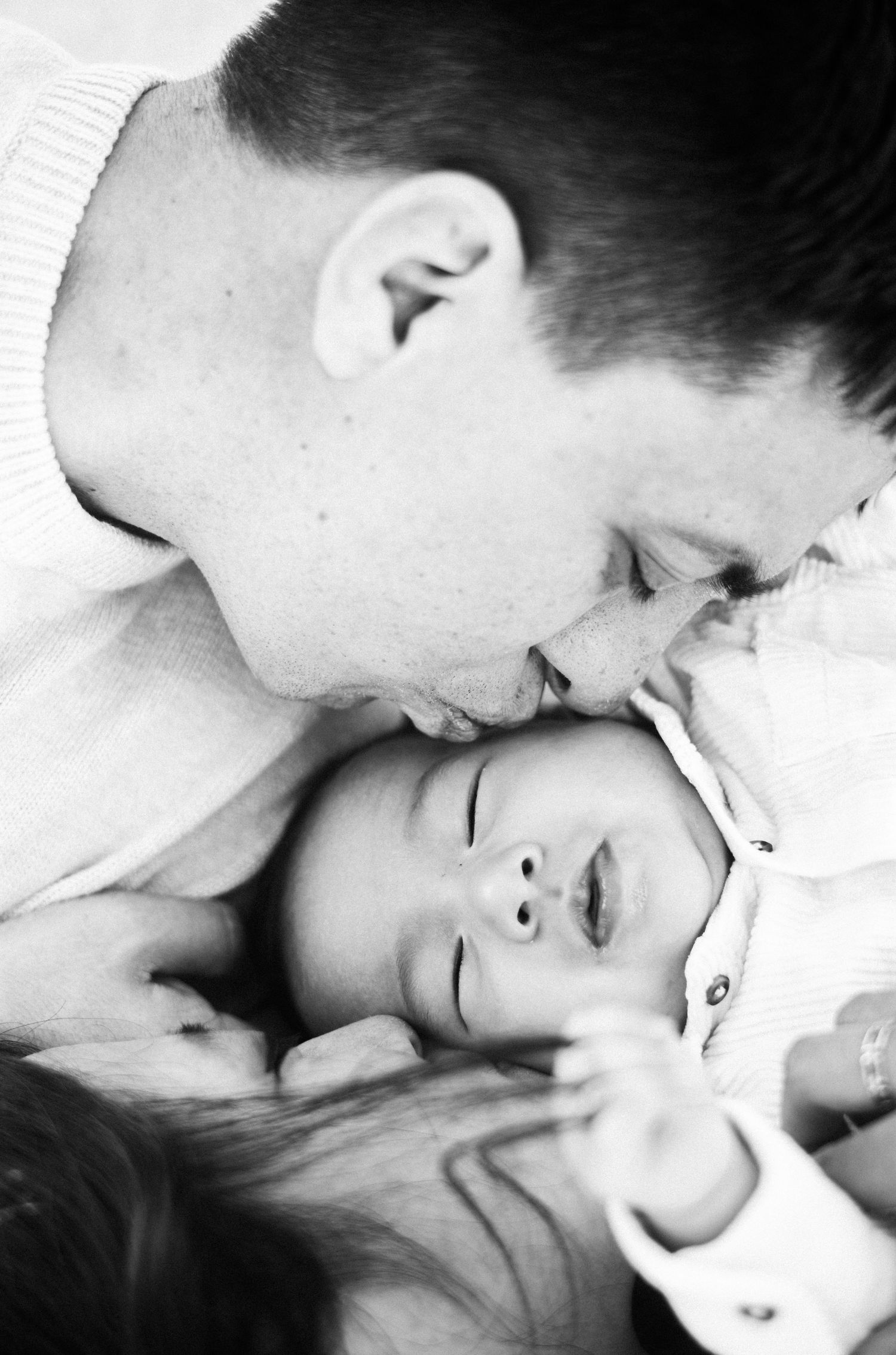 Black and white close-up of a father gently kissing his newborn babys forehead in a tender moment.