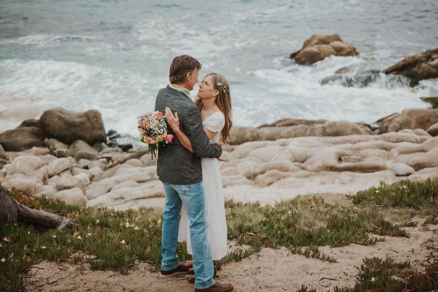 Couple embracing on rocky coastal cliffs, bride in white dress holding bouquet, ocean waves in background.