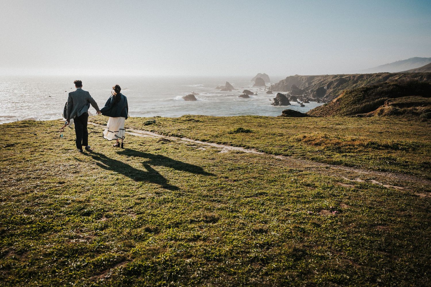 Couple holding hands walking along a scenic coastal cliff path with ocean views and rocky outcroppings.Photography by The Y.K. Studio.