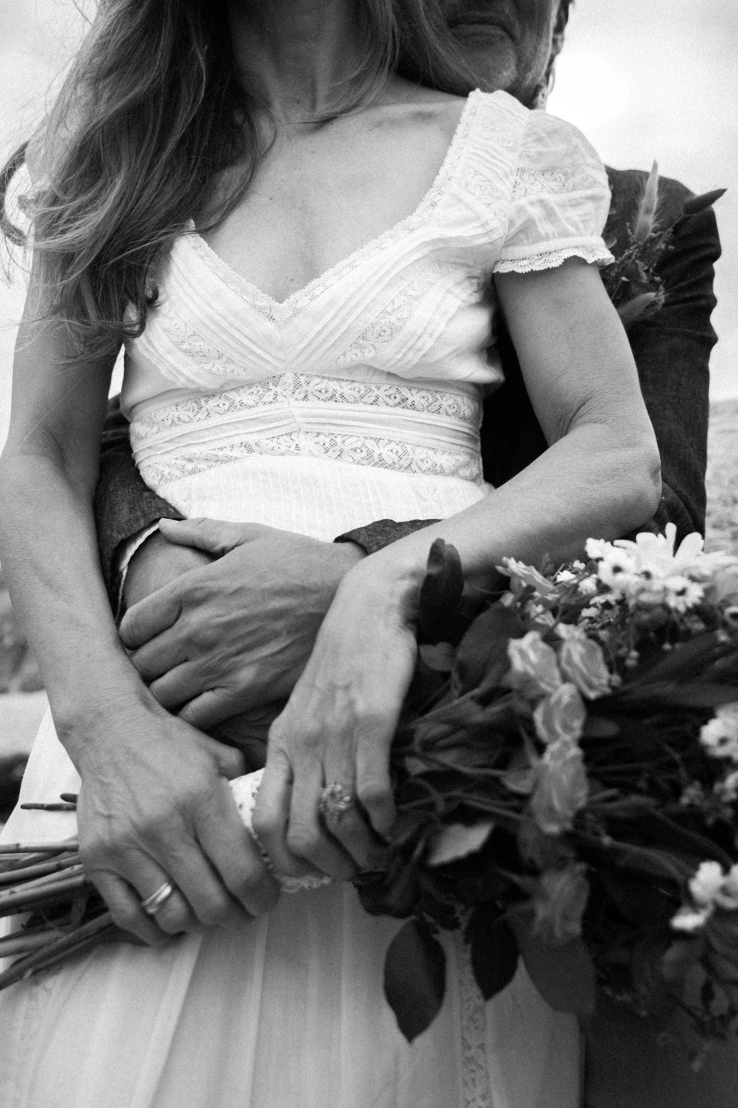 Black and white close-up of bride in lace wedding dress holding floral bouquet with groom embracing her.Photography by The Y.K. Studio.
