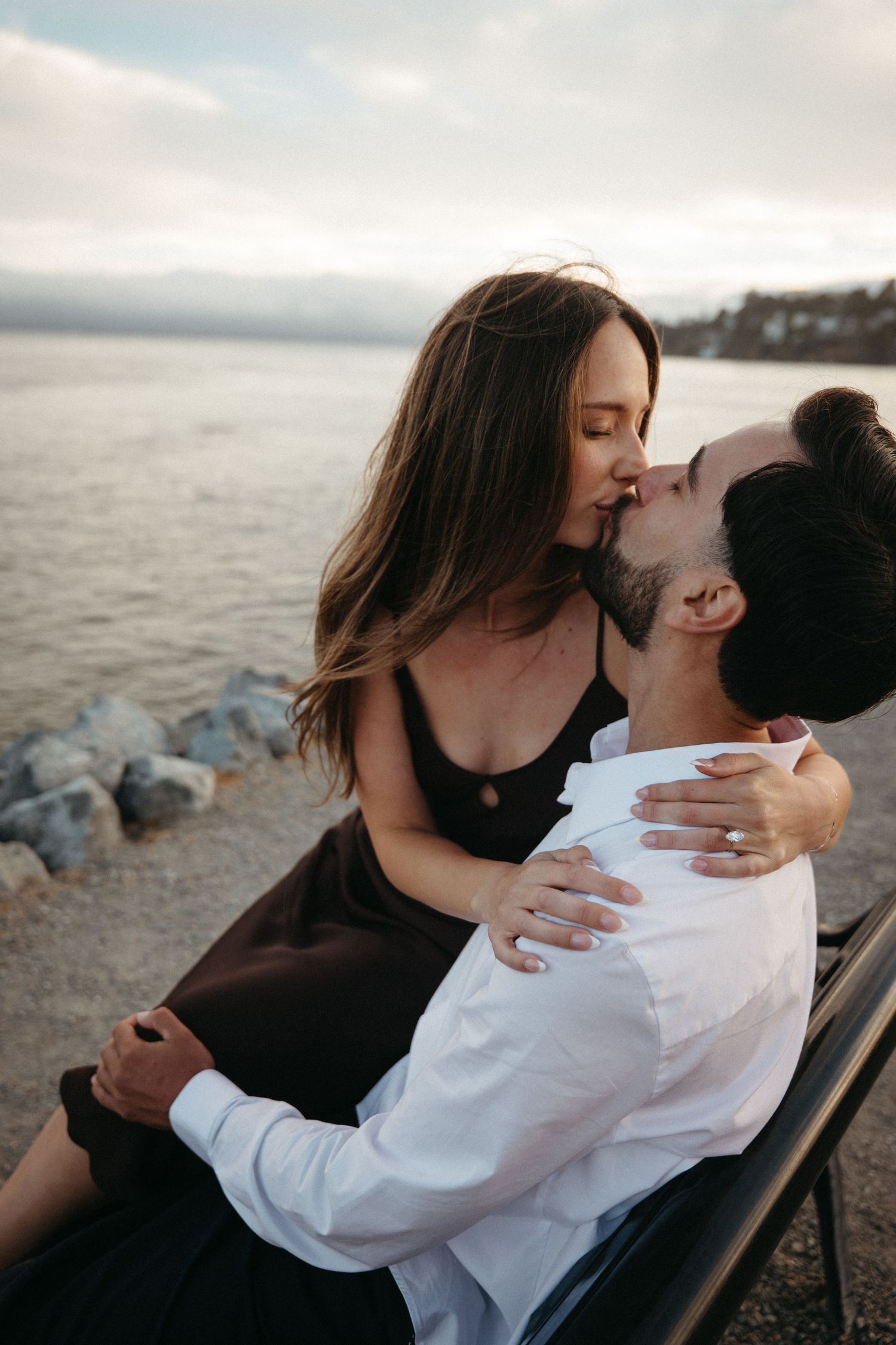 Couple sharing a romantic kiss by the waterfront, woman in black dress sitting on man's lap in white shirt.