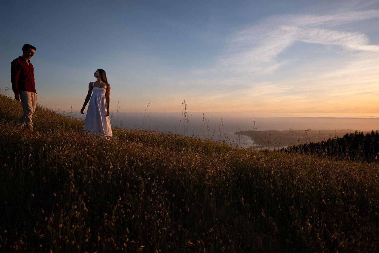 Couple standing on a grassy hillside at sunset with wind turbines and a glowing horizon in the background.