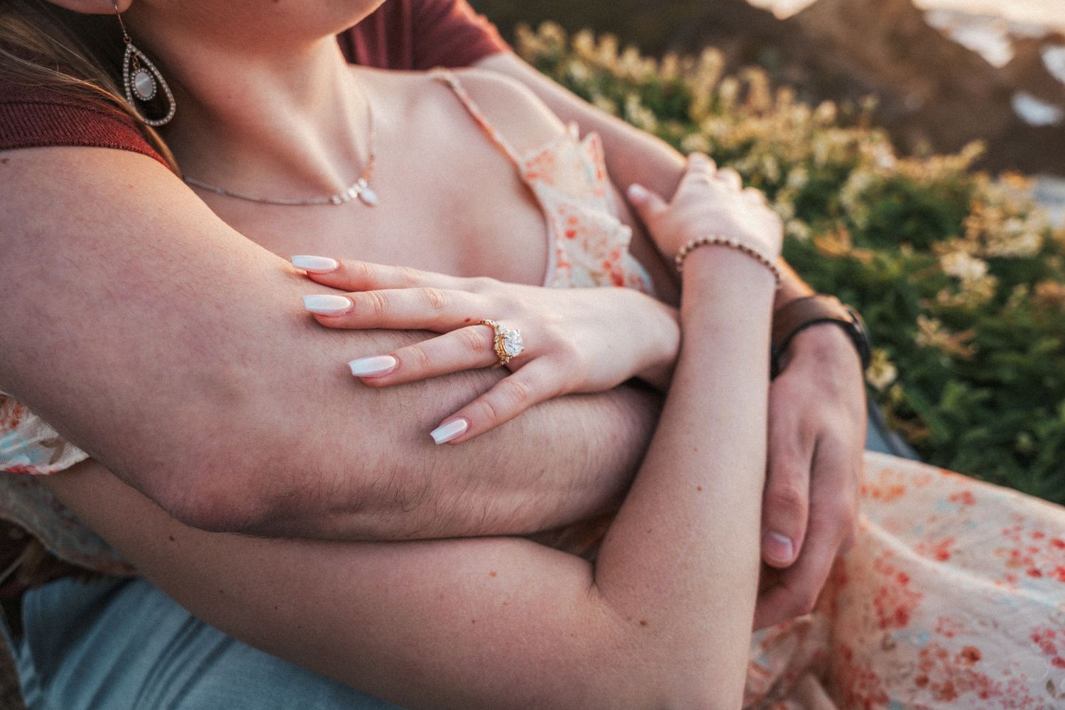 Couple embracing outdoors, woman wearing floral dress, diamond engagement ring, and delicate necklace with greenery background.