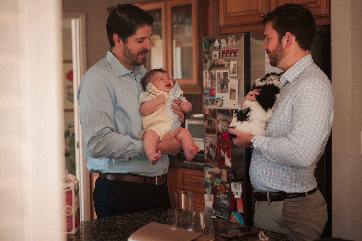 Two men stand in a kitchen, one holding a newborn baby and the other holding a black and white cat.