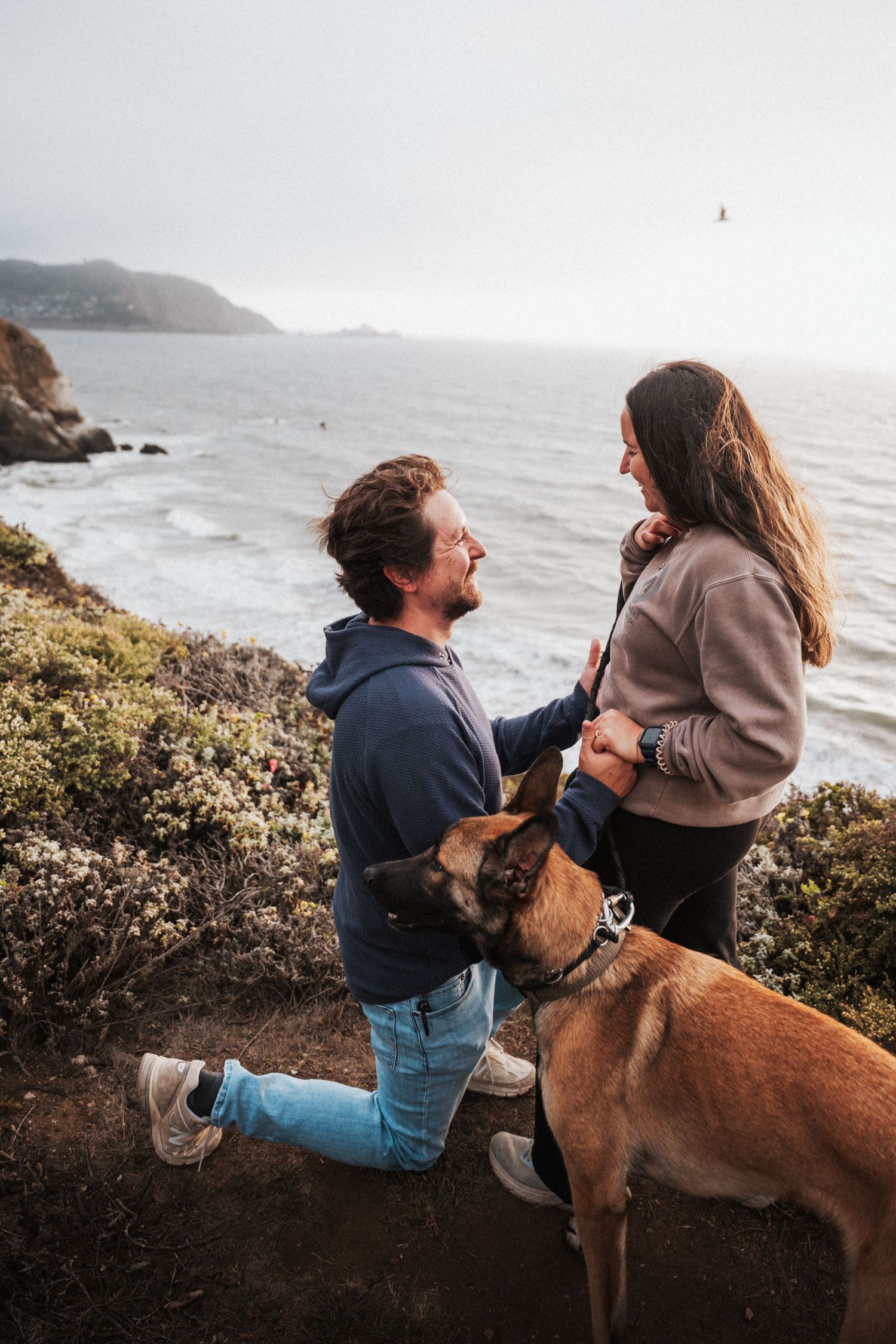 Couple laughing together on coastal cliffside with brown dog and ocean views.