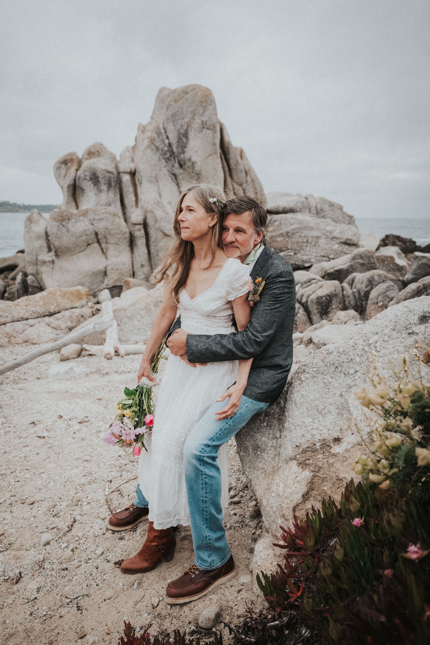 Couple embracing on rocky coastal shore, bride in white dress holding pink bouquet, groom in jeans and gray blazer.