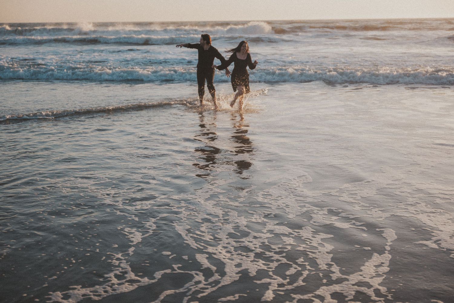 Couple holding hands running through shallow ocean waves on a sandy beach at sunset.Photography by The Y.K. Studio.