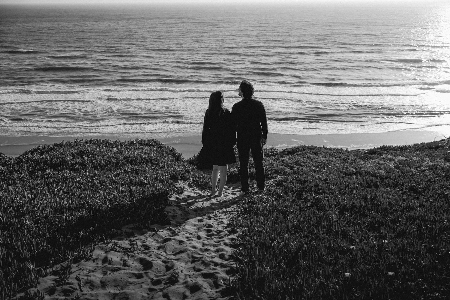 Silhouette of a couple holding hands on a sandy coastal path overlooking the ocean.