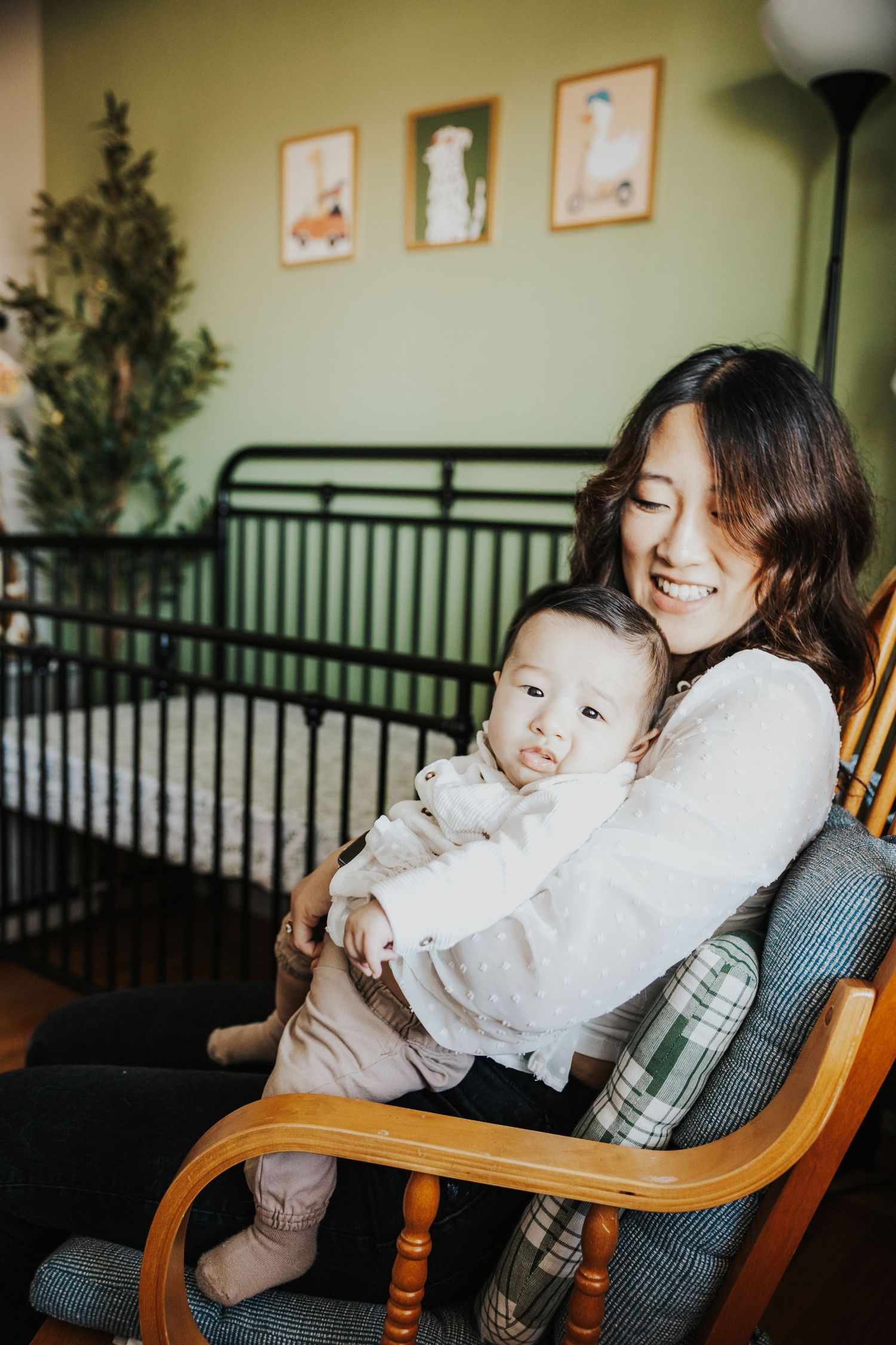 Smiling mother sits in a rocking chair holding her happy baby in a cozy green nursery room.