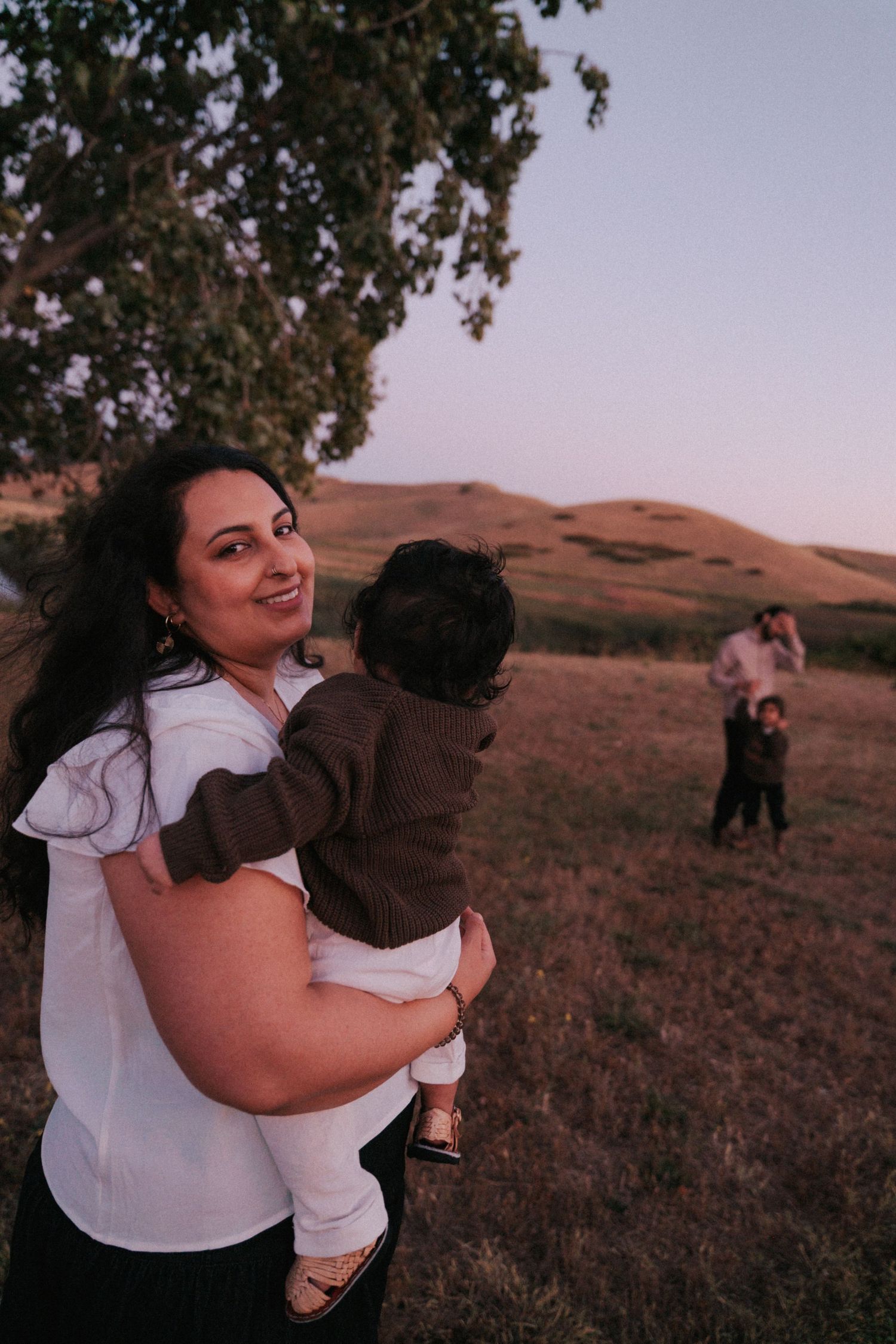 A woman smiles while holding a baby outdoors at dusk, with rolling hills and a couple in the background.