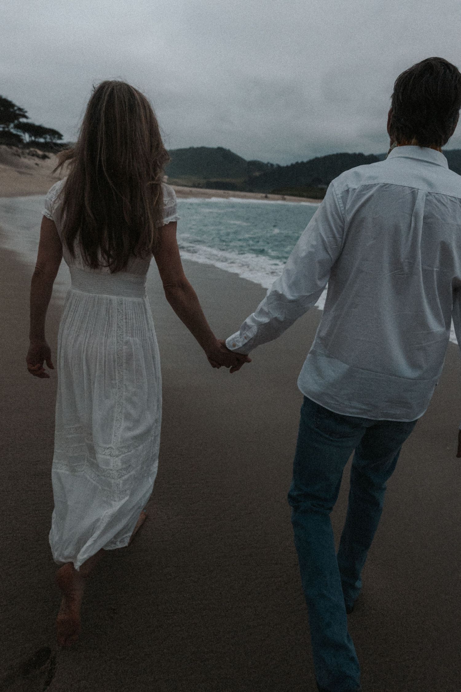 Couple holding hands walking along a moody beach, woman in white dress, man in white shirt and jeans.Photography by The Y.K. Studio.