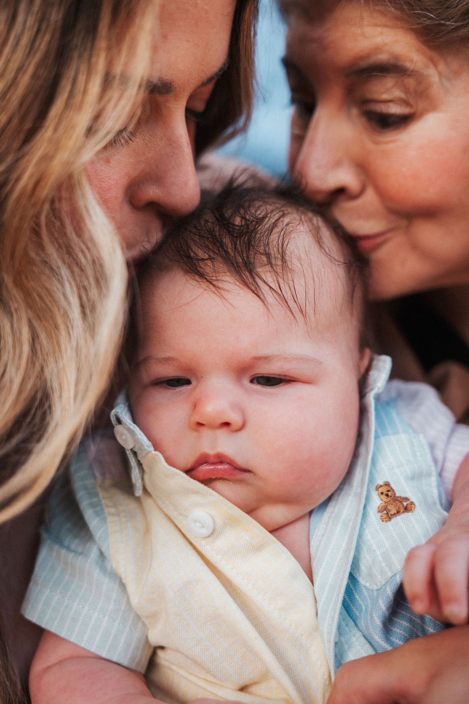 Two women lovingly kiss a newborn baby dressed in a light blue striped outfit with a teddy bear pin.