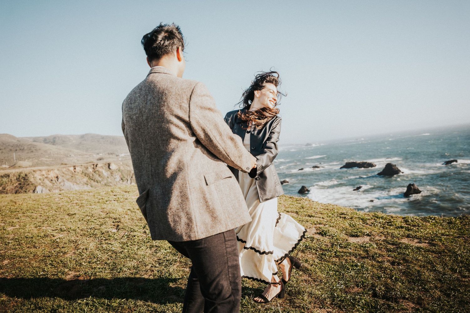 Couple holding hands on a coastal cliff, woman's hair blowing in the wind, ocean backdrop.