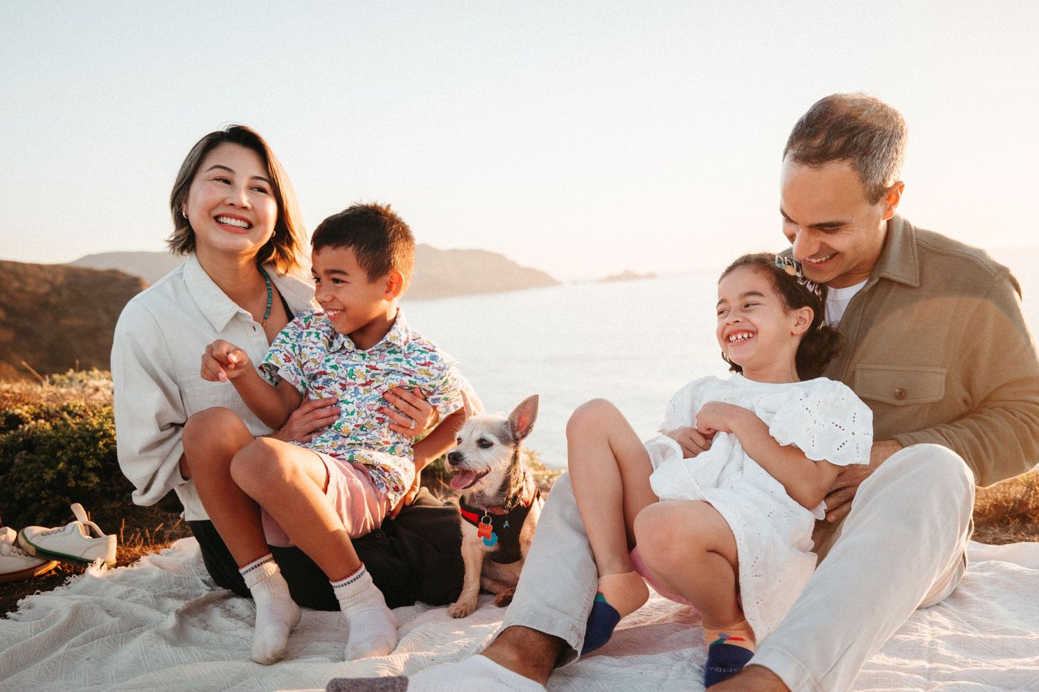 Happy family with two kids and a small dog enjoying a sunny beach picnic by the ocean at sunset.