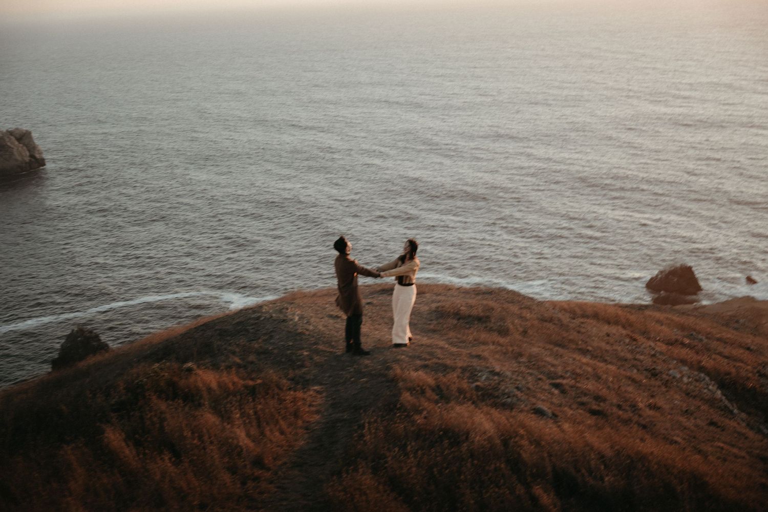 Couple holding hands on a grassy coastal cliff overlooking the ocean at sunset during an intimate elopement.