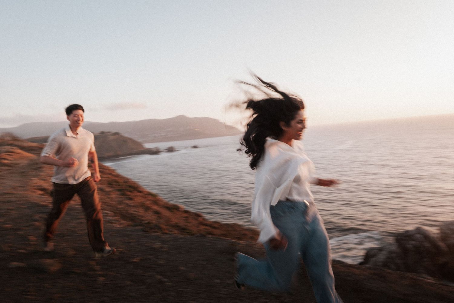 Couple running along a coastal cliff at sunset, woman's hair blowing in the wind with ocean backdrop.