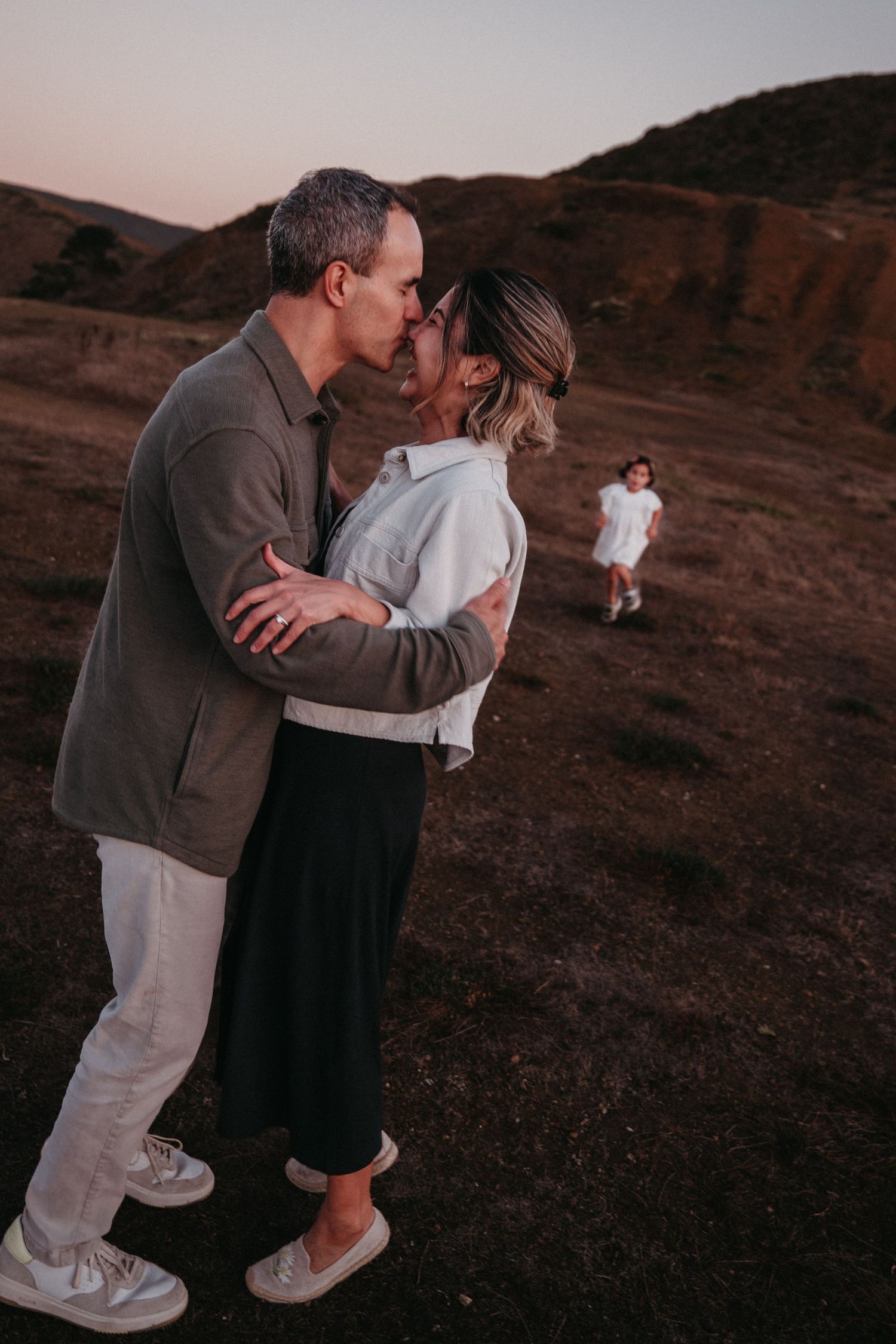 A couple shares a romantic kiss on a hillside at dusk with a child in the background.