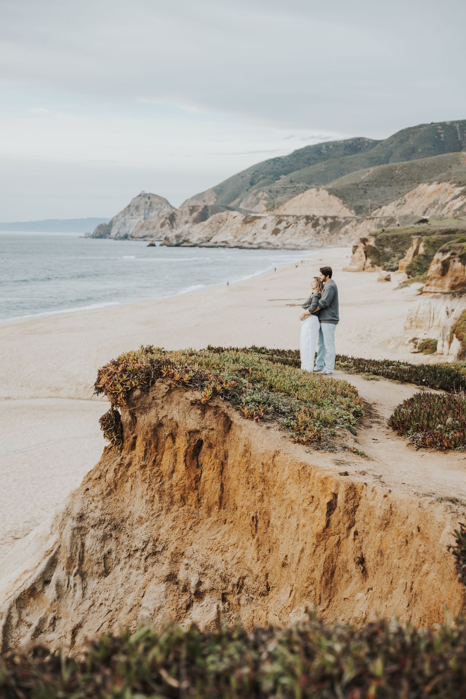 Person standing on coastal cliffs overlooking a sandy beach with green hills and ocean in the background.