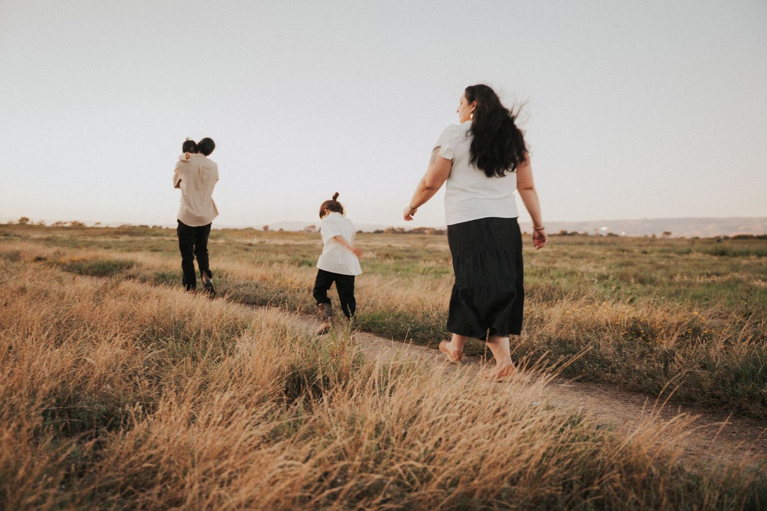 Family walking on a dirt path through golden grasslands at dusk, mother and child holding hands outdoors.