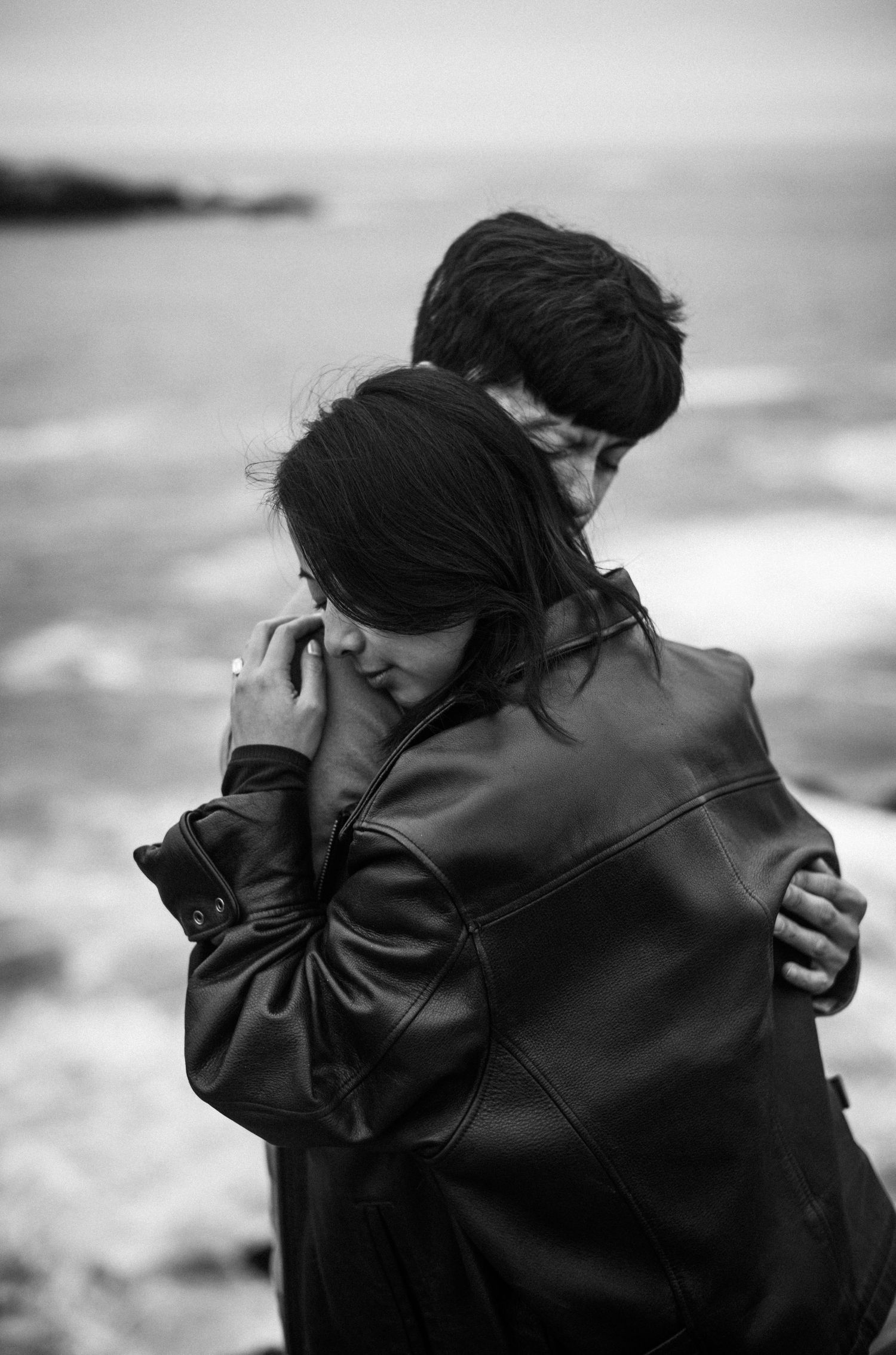 Emotional black and white photo of couple embracing tightly near the ocean, woman hiding her face.
