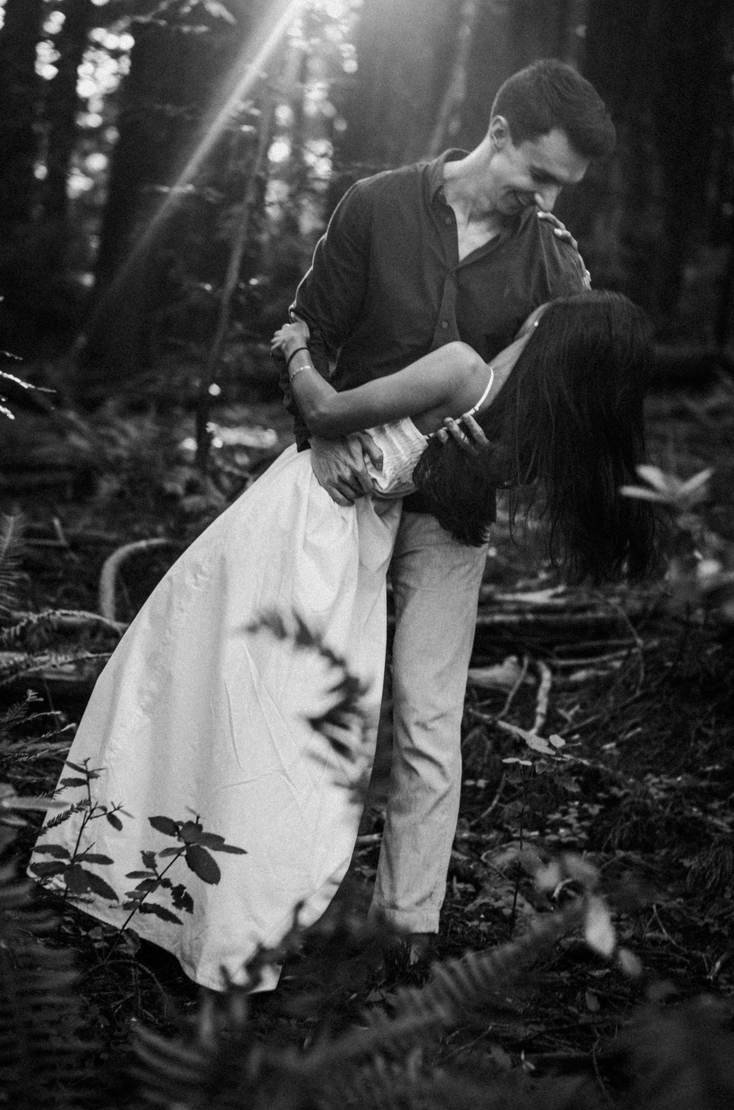 Black and white photo of a couple romantically embracing and dipping in a sunlit forest setting.