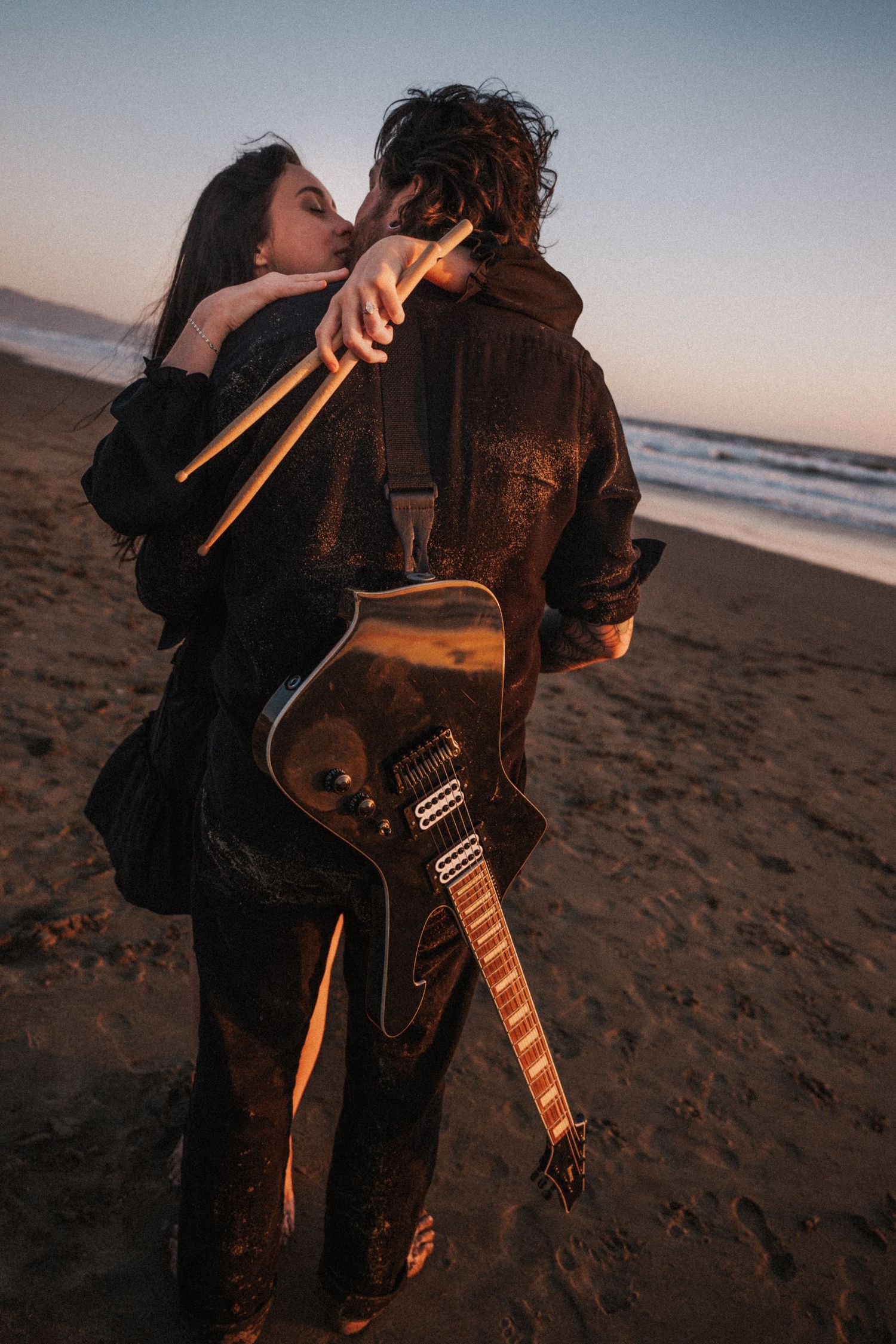 Couple embracing and kissing on a beach at sunset, man holding an electric guitar.
