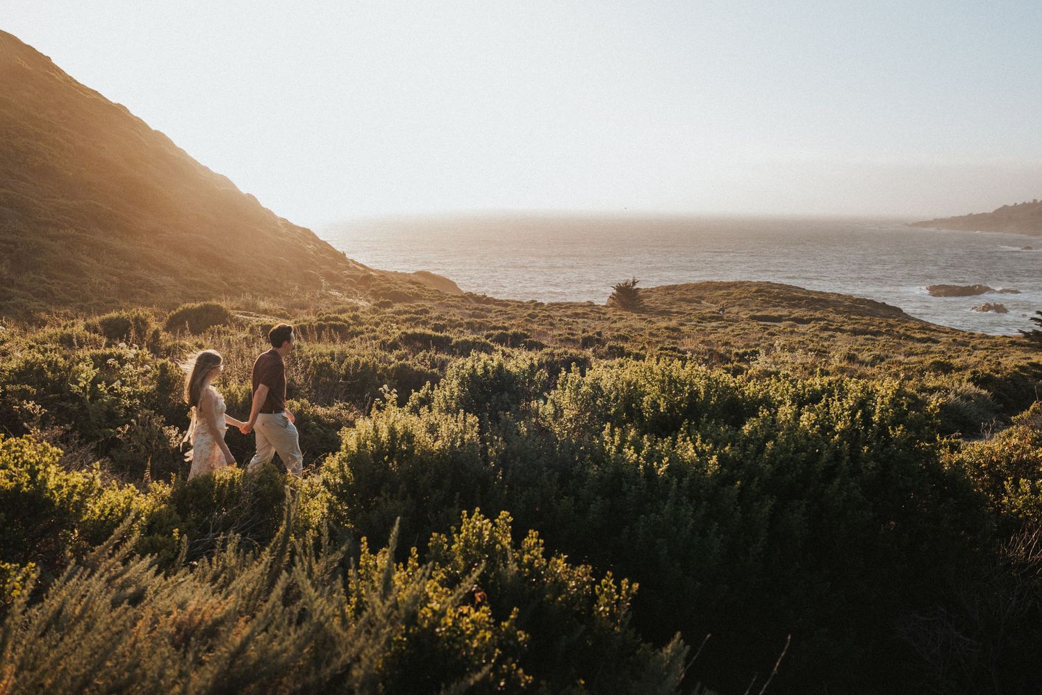 Couple walking through golden coastal shrublands at sunset with ocean views in Big Sur, California.