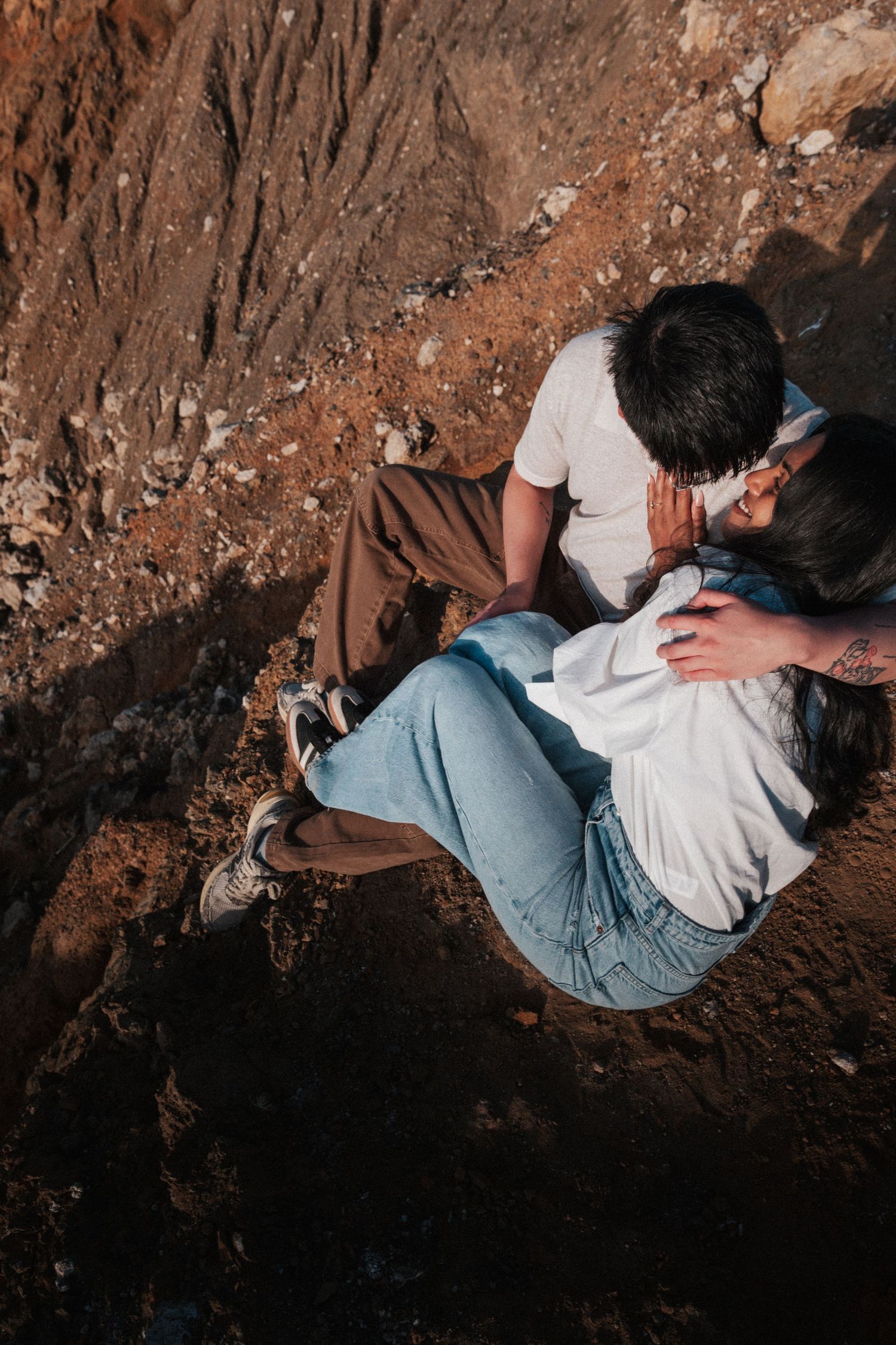Couple sitting together on rocky terrain, embracing romantically, wearing casual white tops and jeans, viewed from above.