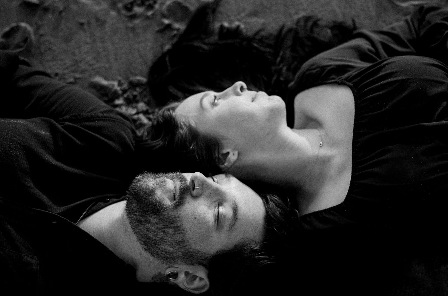 Black and white photo of two people lying on the ground with eyes closed, wearing black clothing.