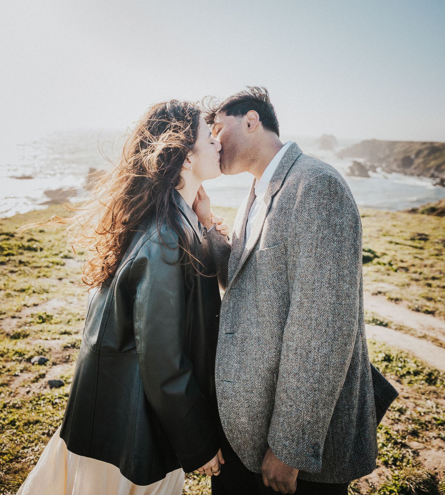 Couple sharing a romantic moment on a scenic coastal cliff, woman in leather jacket and man in grey tweed coat.