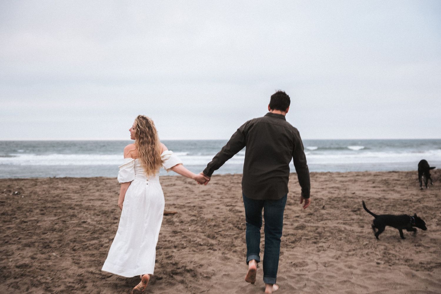 Couple holding hands walking on sandy beach toward ocean waves with a black dog nearby.