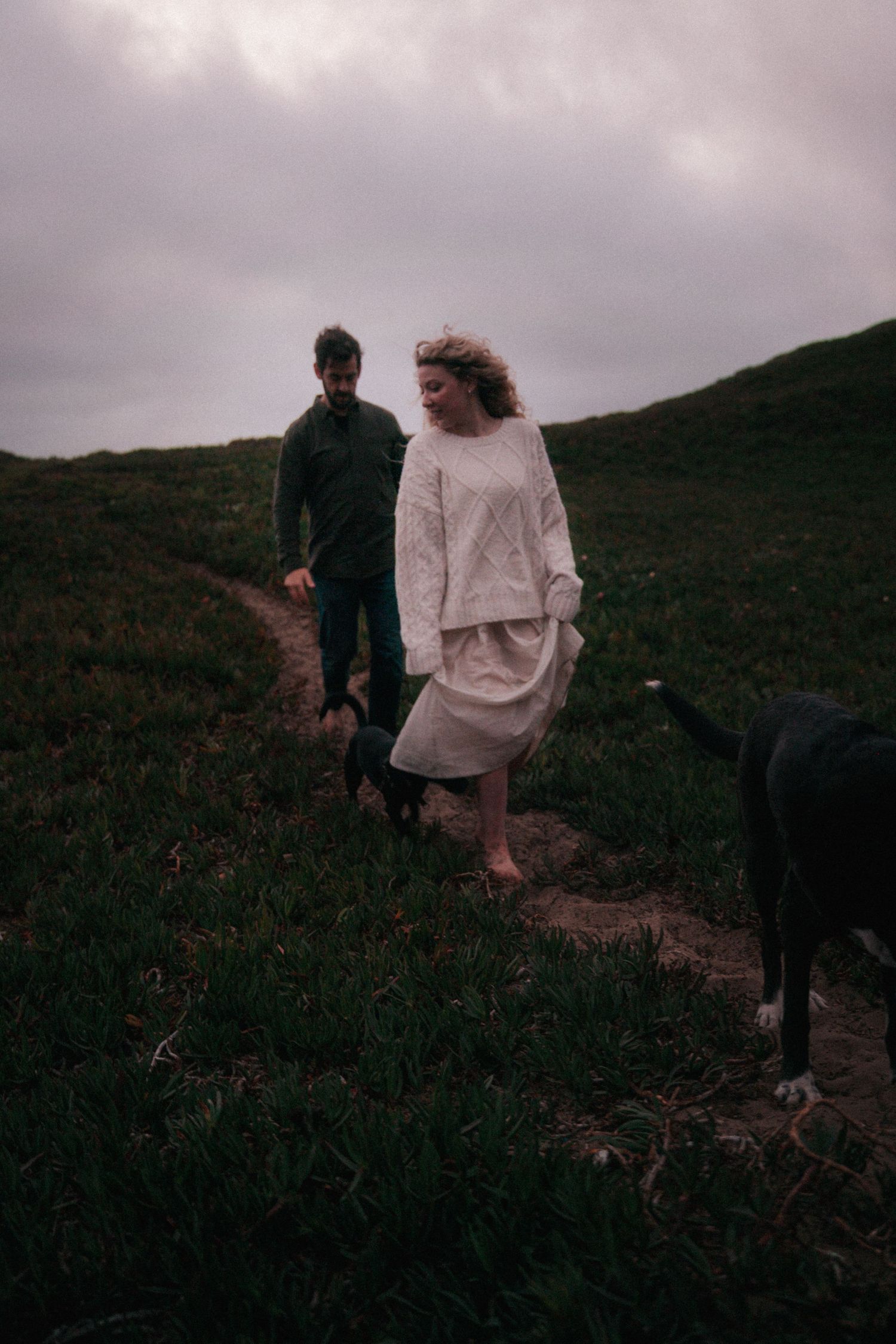 Couple walking barefoot on a grassy hillside path with two black dogs under a cloudy sky.