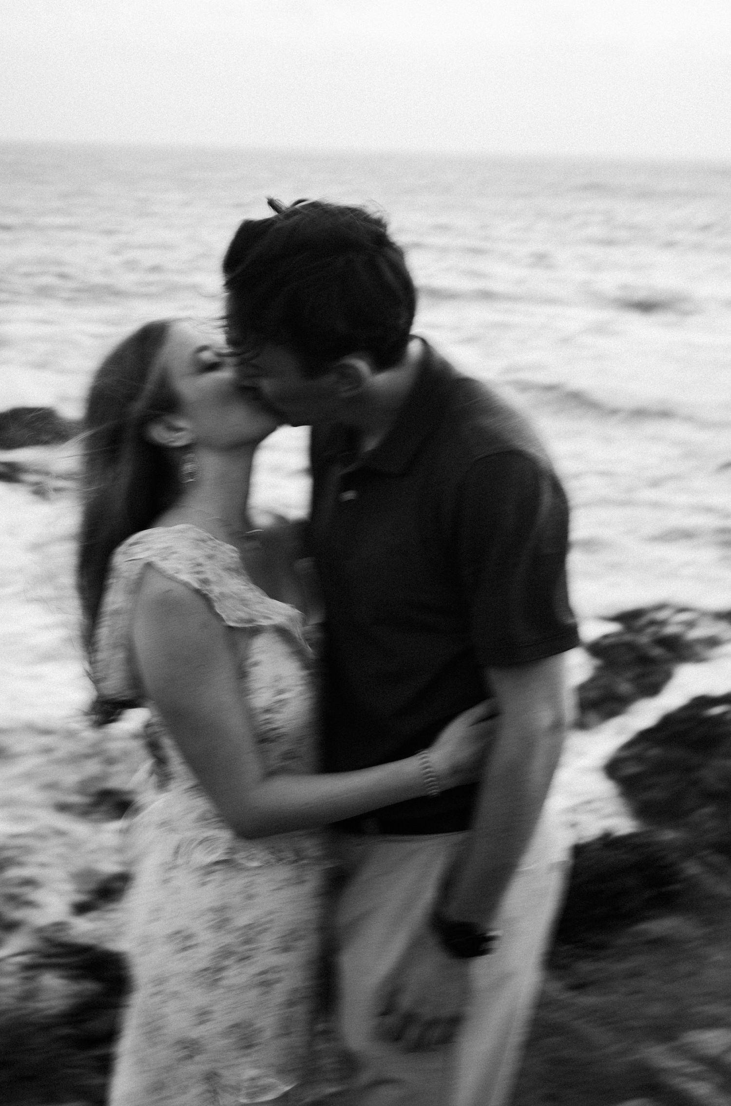 Black and white blurred artistic photo of couple kissing on rocky ocean shoreline.