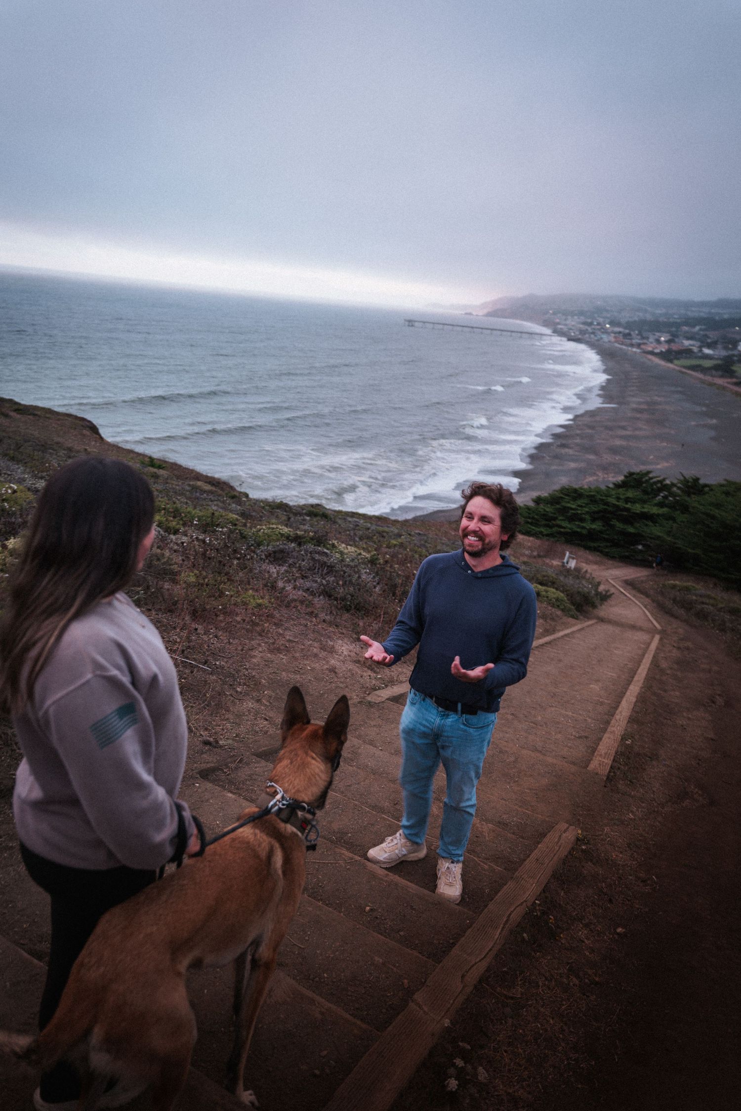 Couple with Belgian Malinois dog on coastal overlook trail with foggy beach and ocean view.