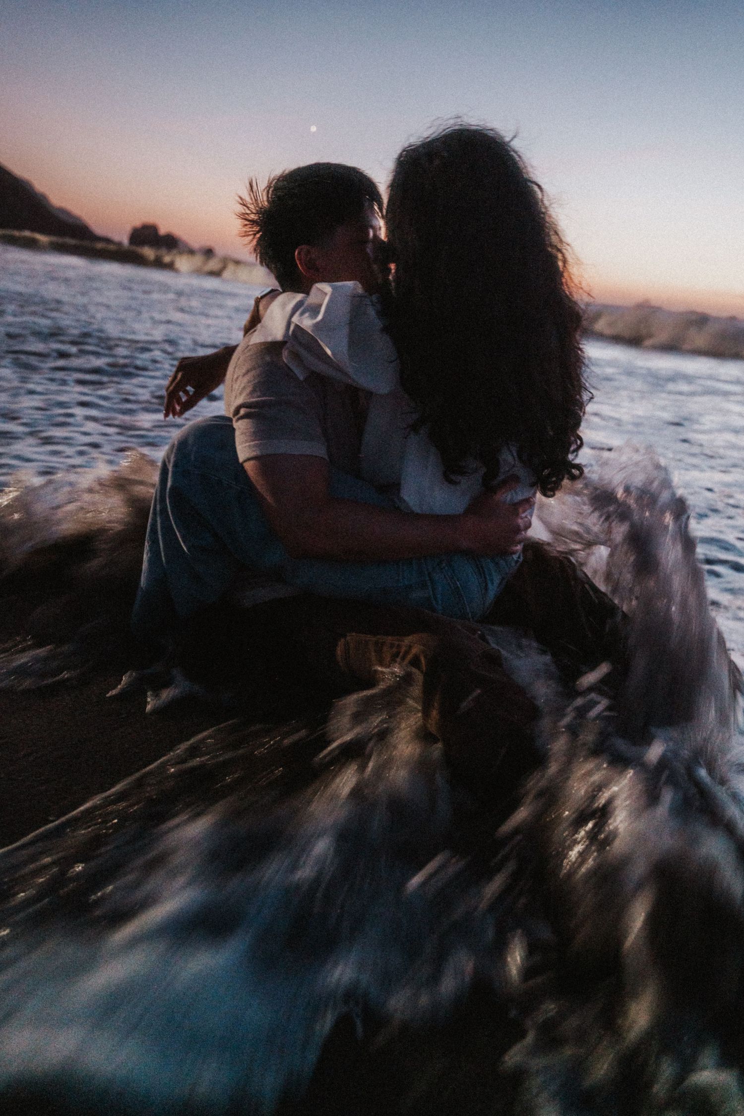 Couple sharing an intimate kiss while sitting together in crashing ocean waves during a colorful sunset.