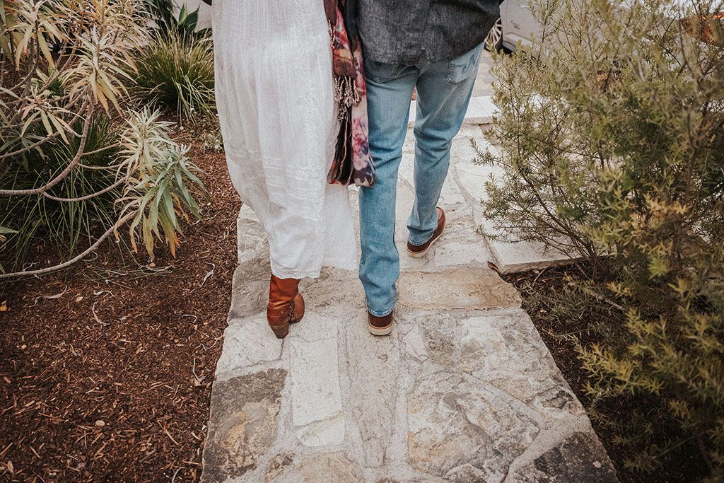 Couple walking on stone path in garden, woman in white dress and man in jeans with cowboy boots.