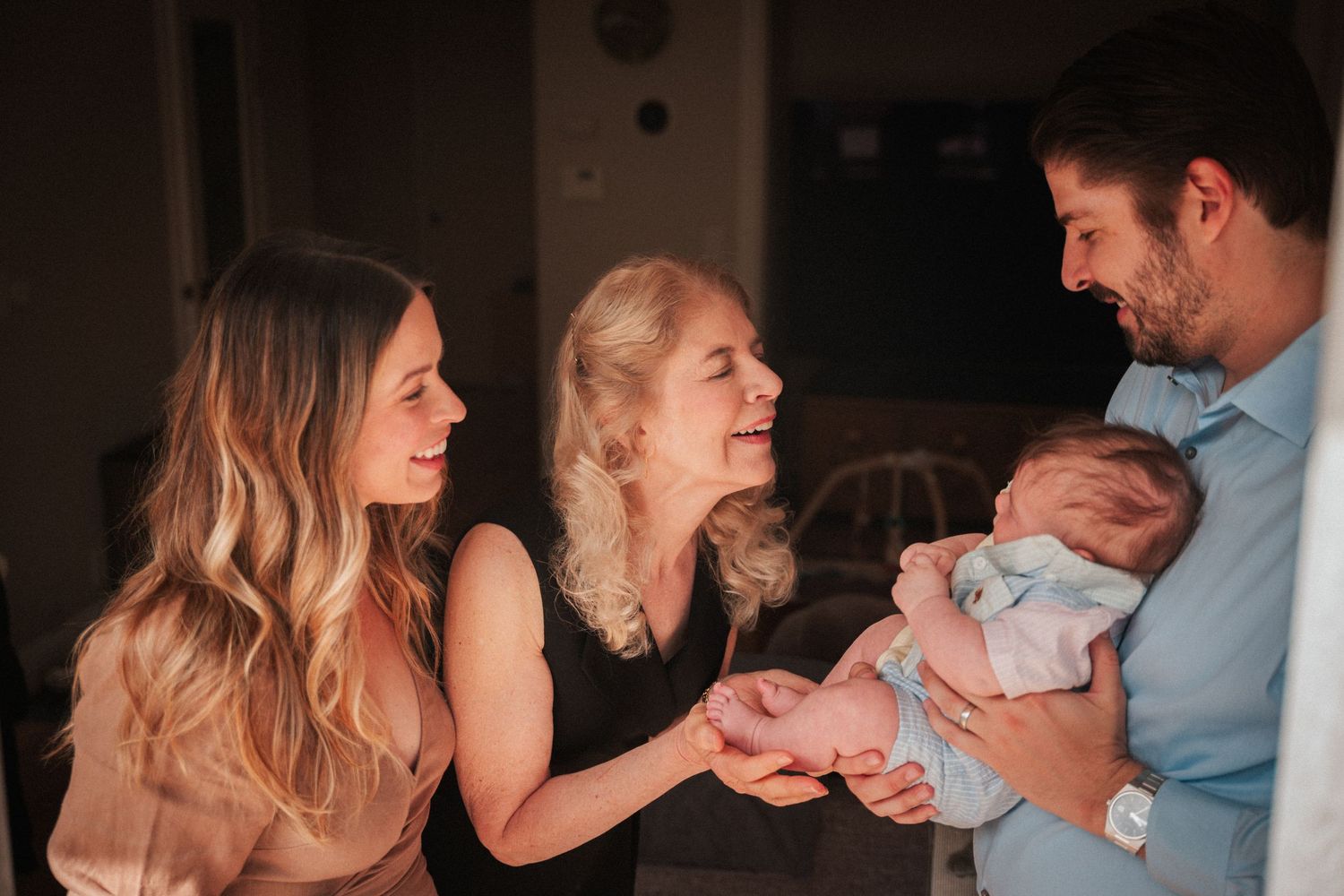 Family gathering as two women admire a newborn baby held by a man in a warmly lit home setting.