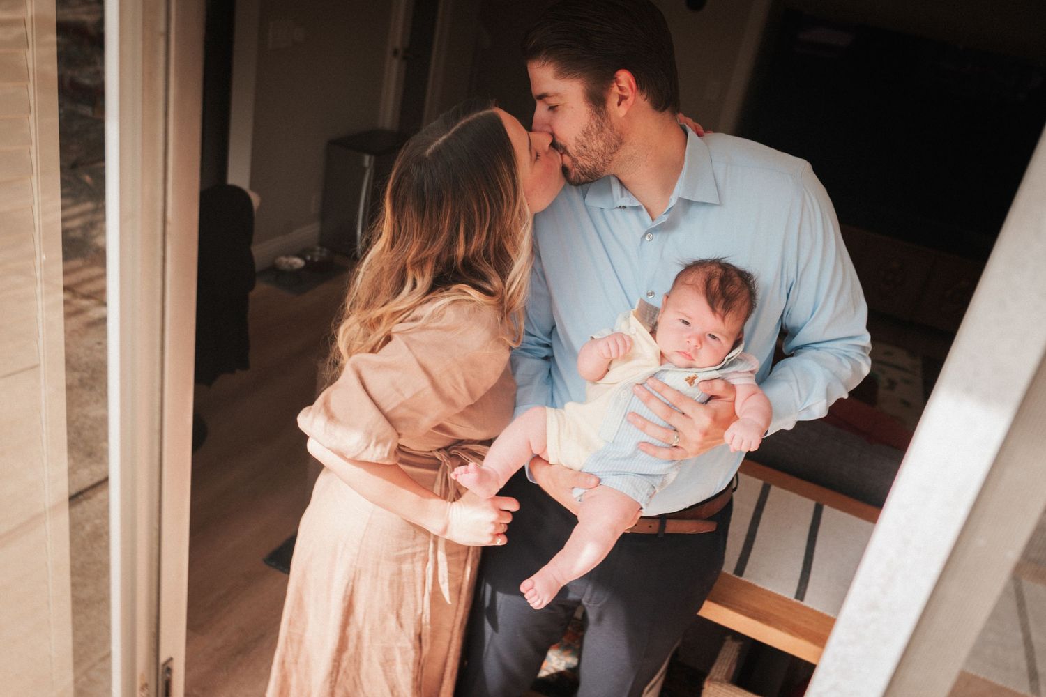 A couple shares a tender kiss while holding their newborn baby in a warmly lit home setting.