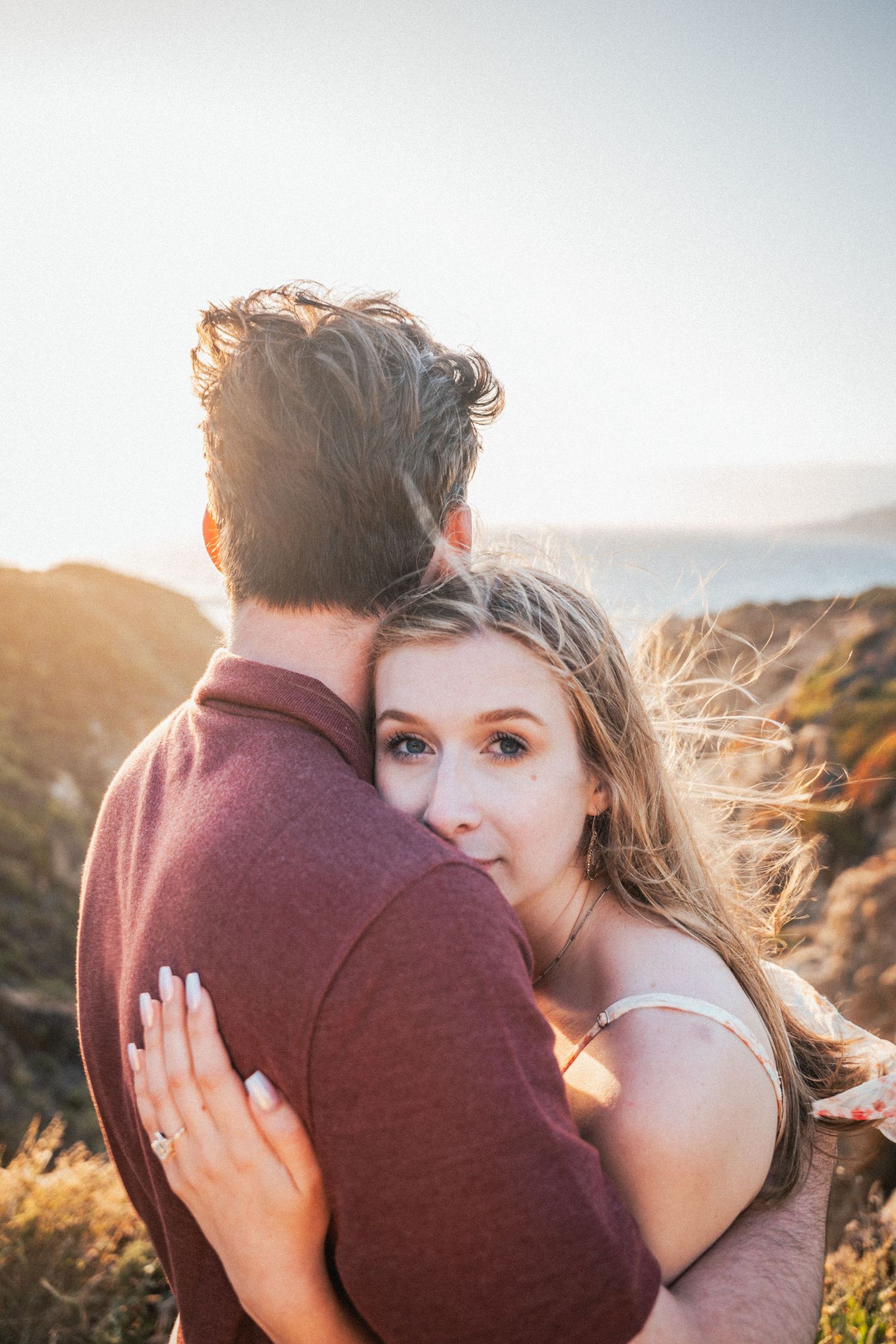 Couple embracing at golden hour on a coastal cliff, woman looking back at camera with wind-blown hair.